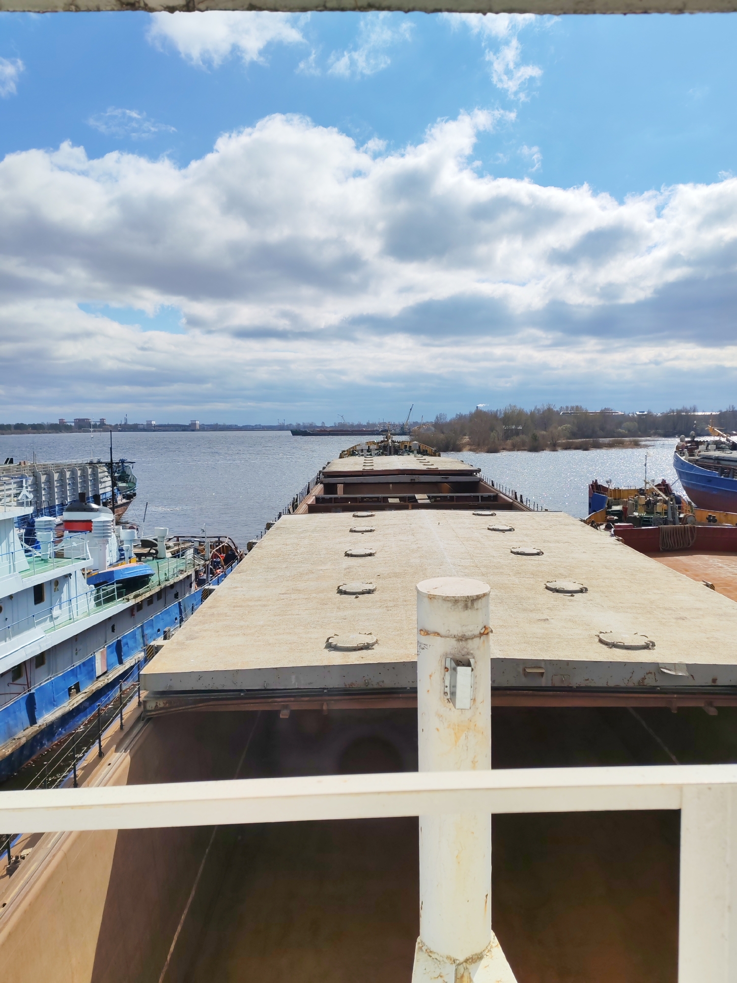 The image shows a view from a ship in Rybinsk, Russia. In the foreground is part of the ship the photo is taken from: white railings, a white post, and a view into the hull of the ship. Beyond that is another, longer, flat-topped barge with circular access points on the top. To the left of the barge is another ship with a light blue hull and various deck fixtures. To the right of the barge, another ship is partially visible. In the background, a wide river flows, reflecting sunlight. On the far bank of the river, there are buildings, trees, and possibly industrial structures visible. The sky is blue with numerous puffy white clouds.