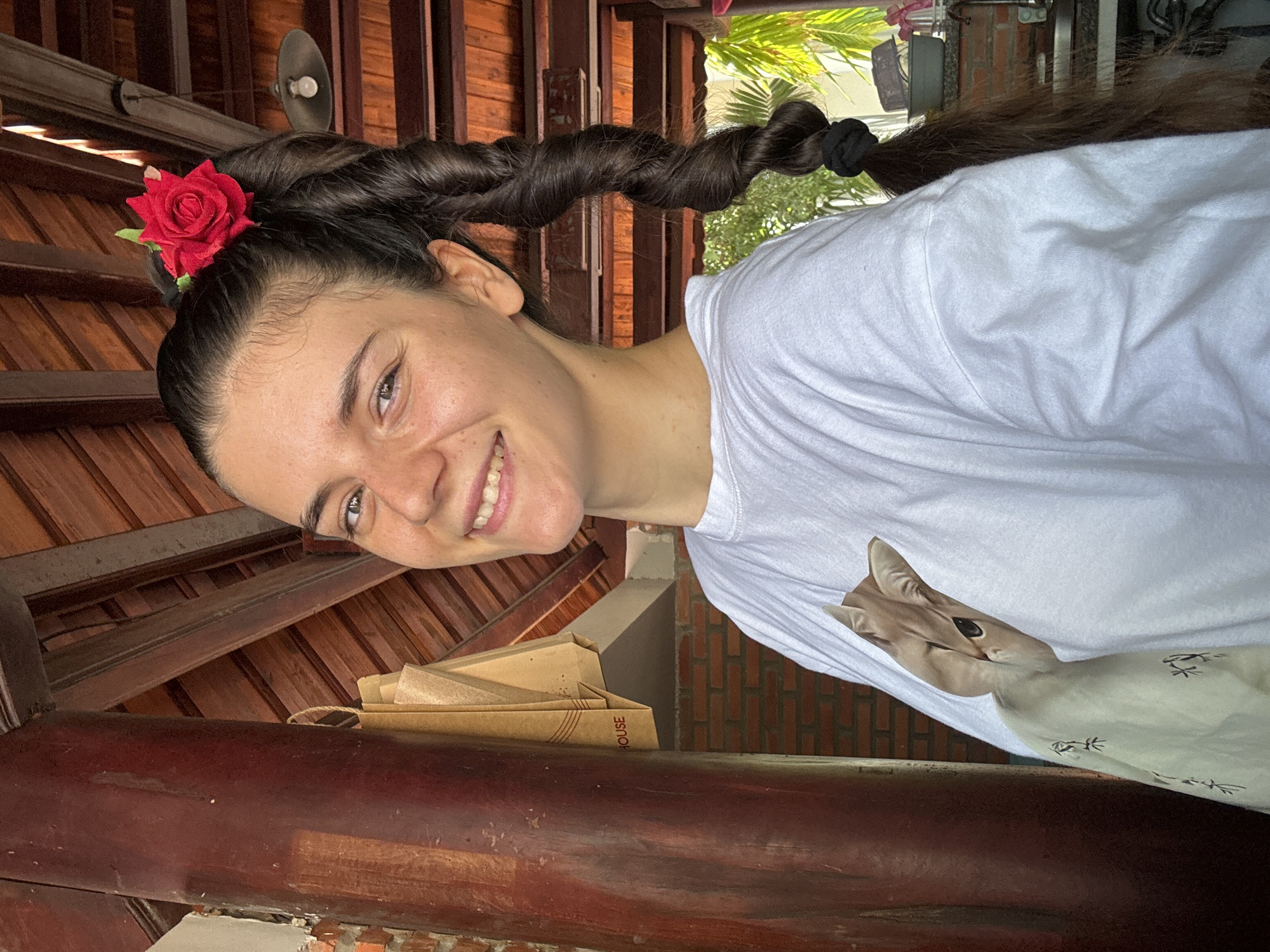 A woman smiles towards the camera, her head adorned with a red flower. Her dark hair is twisted into a thick braid. She wears a white t-shirt imprinted with an image of a cat. The background suggests a rustic, partially-covered outdoor setting. Wooden beams form a ceiling structure, while foliage is visible outside. Stacks of brown paper bags are present. The brick wall in the background suggests a building material common in Vietnam. Given the architectural style and background elements, the location is likely within Mũi Né, Vietnam.