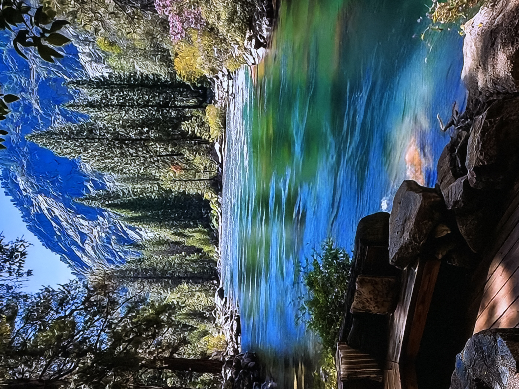 A landscape photograph, oriented sideways (rotated 90 degrees counter-clockwise), captures a serene natural scene. The primary subject is a vibrant, clear river with striking blue and green hues, reflecting the surrounding environment.
On the left side (when viewed correctly), majestic, rugged mountains rise into a bright blue sky, their surfaces showing textured patterns. Dense coniferous forests, primarily dark green pine or fir trees, line the riverbanks and ascend the mountain slopes, creating a rich green canopy. Interspersed among the darker evergreens, some trees display lighter green, yellowish, and even pinkish-purple foliage, suggesting diverse plant life or a specific season like spring or early autumn.
In the foreground, on the right side, there is a partially visible man-made structure composed of rough-hewn wooden beams and large, natural-looking stones. This element, possibly part of a viewing platform, bridge, or cabin, blends into the rocky bank of the river. Small leafy green bushes grow near this structure and along the river's edge.
The scene depicts a calm, sunlit day with clear weather, evoking a sense of untouched natural beauty and tranquility. There is no specific activity taking place, and no visible text is present in the image.
