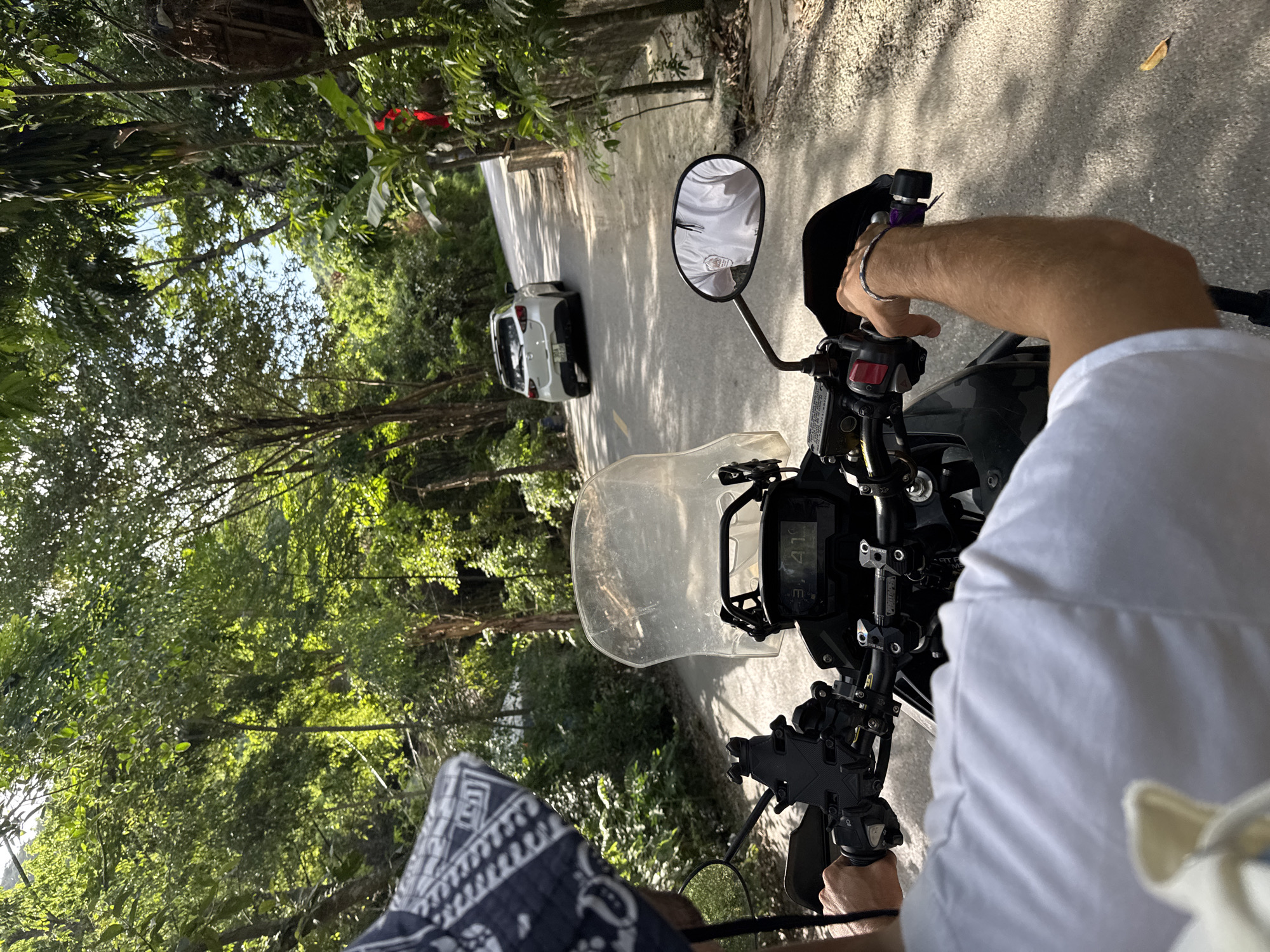 A first-person perspective shot from a motorcycle shows a paved road winding through a lush, green forest. Sunlight filters through the dense canopy, casting dappled shadows on the asphalt. The road ahead is clear, with a white car visible in the distance, also traveling forward. The motorcycle's handlebars and instrument panel are in focus, with the rider's arm and hand resting on the controls. The rider is wearing a white t-shirt and a patterned bandana on their head. The surrounding vegetation is rich and varied, with large trees and smaller plants lining both sides of the road. The overall impression is one of a scenic ride through a tropical or subtropical environment, possibly within the city of Nam Hoa Lư Ward, Vietnam.