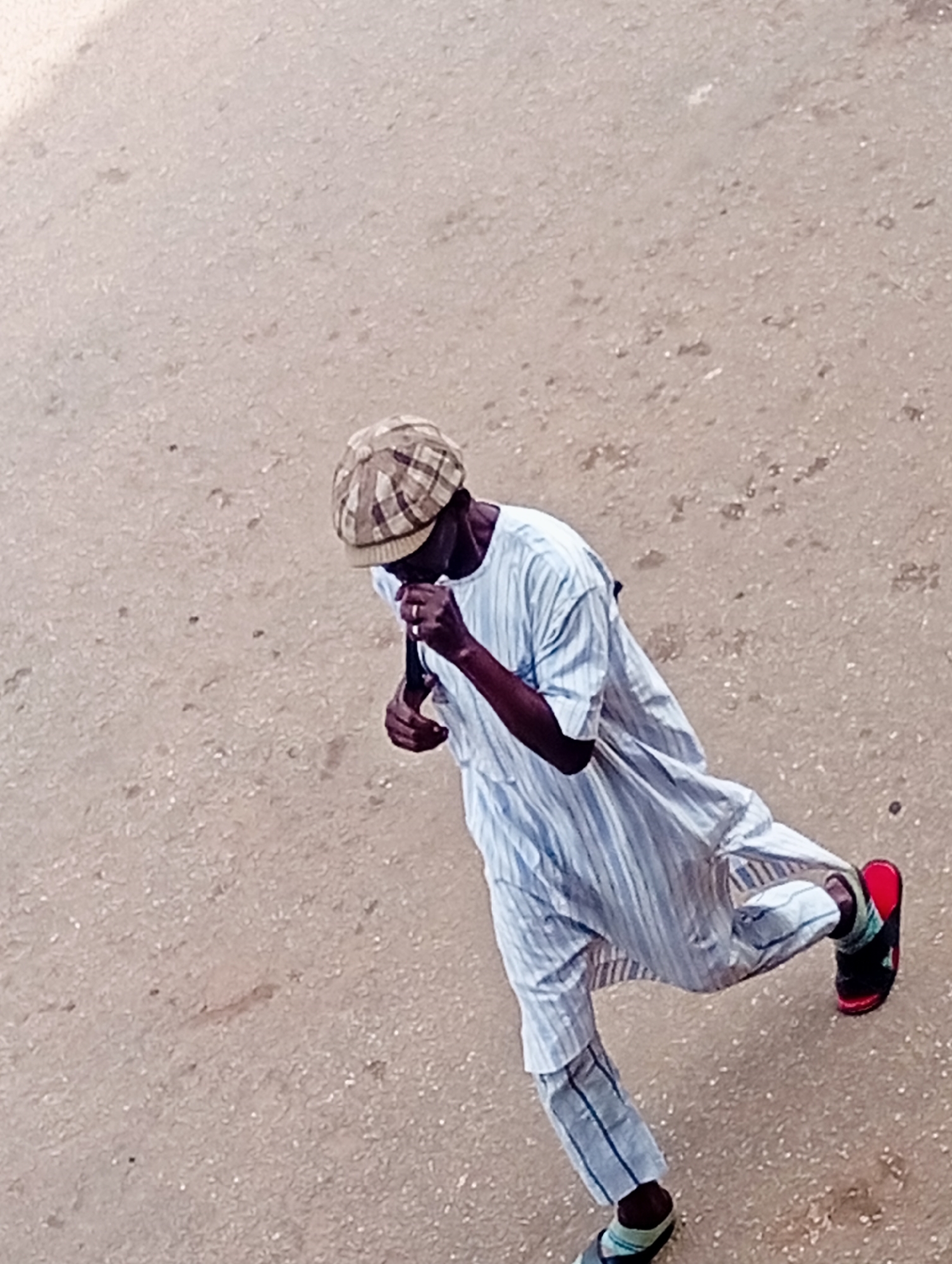 In Bukuru, Nigeria, a person wearing a plaid cap and a light blue striped garment is captured from a high angle as they walk on a sandy or dirt surface. The individual's hands are near their face, suggesting they might be adjusting their clothing or holding an object. They are wearing striped trousers and sandals, one of which is red. The ground is a textured brown, with scattered small particles.