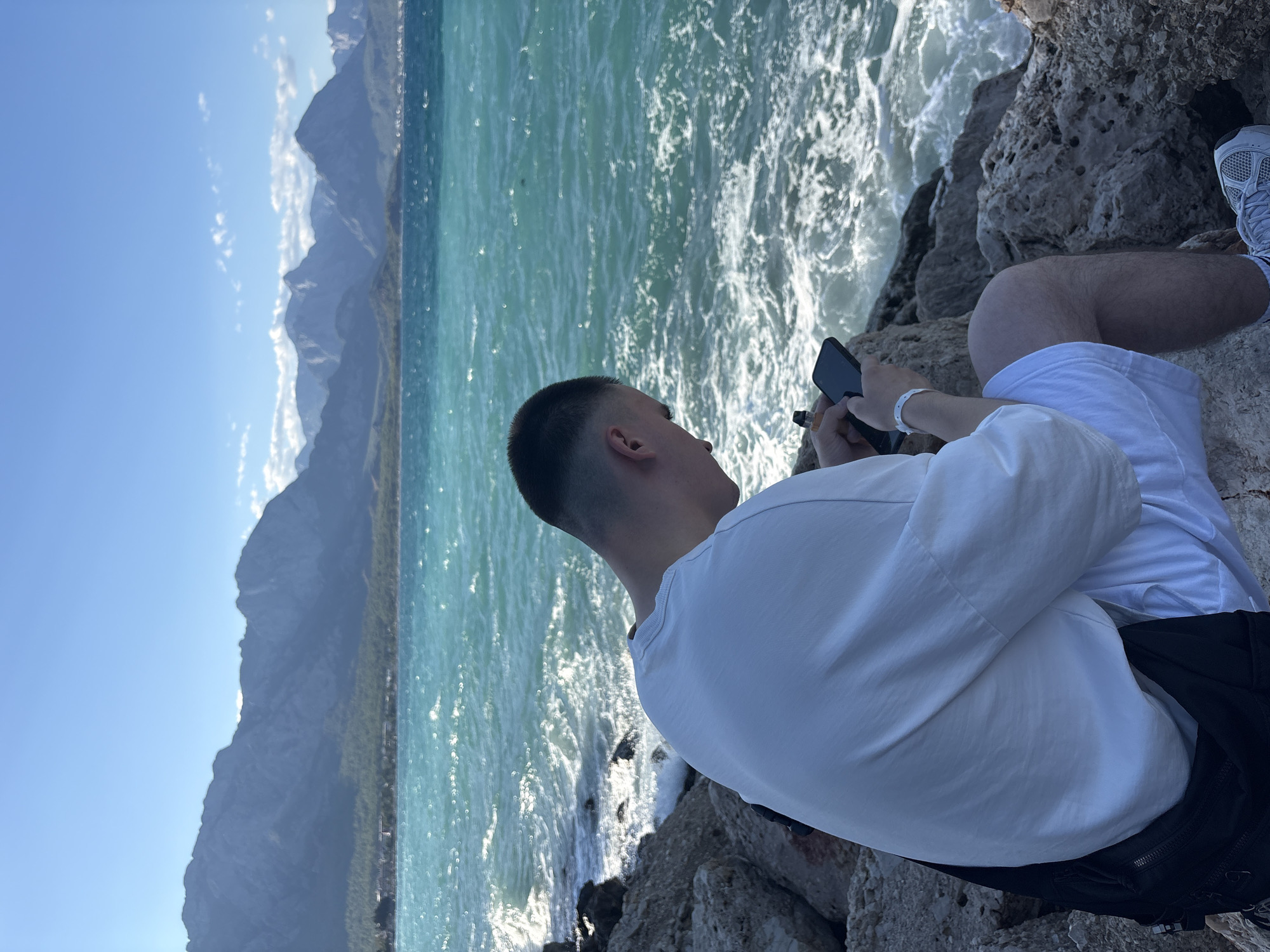 A young man with a fade haircut, wearing a white t-shirt and white shorts, sits on a rocky shoreline overlooking the turquoise waters of the Mediterranean Sea. Mountains form a distant backdrop, under a clear blue sky dotted with a few clouds. The man is looking down at his phone, and a black bag is slung over his shoulder. The waves are breaking on the rocks, creating a foamy white splash. The scene appears to be near Kemer, Turkey, a popular coastal resort town known for its beaches and proximity to the Taurus Mountains.