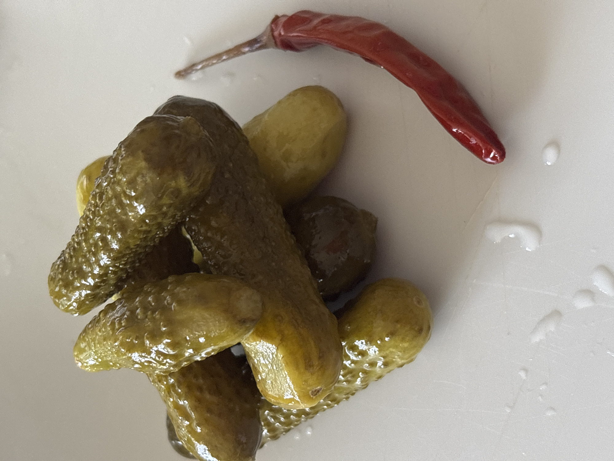 This is a close-up, eye-level, medium shot of a pile of gherkins and a dried red chili pepper on a white surface. The gherkins are green, bumpy, and appear to be pickled. They are stacked haphazardly on top of each other, with some appearing to be partially submerged in liquid. The dried chili pepper is long, slender, and deep red, lying to the right of the gherkins. The white surface appears to have some droplets of liquid on it, suggesting it might be a plate or countertop. The lighting is bright and natural, casting subtle shadows. There are no people or animals in the image. The background is out of focus and a neutral, light color. This image is not specific to the city of Sochi, Russia, and could have been taken anywhere.