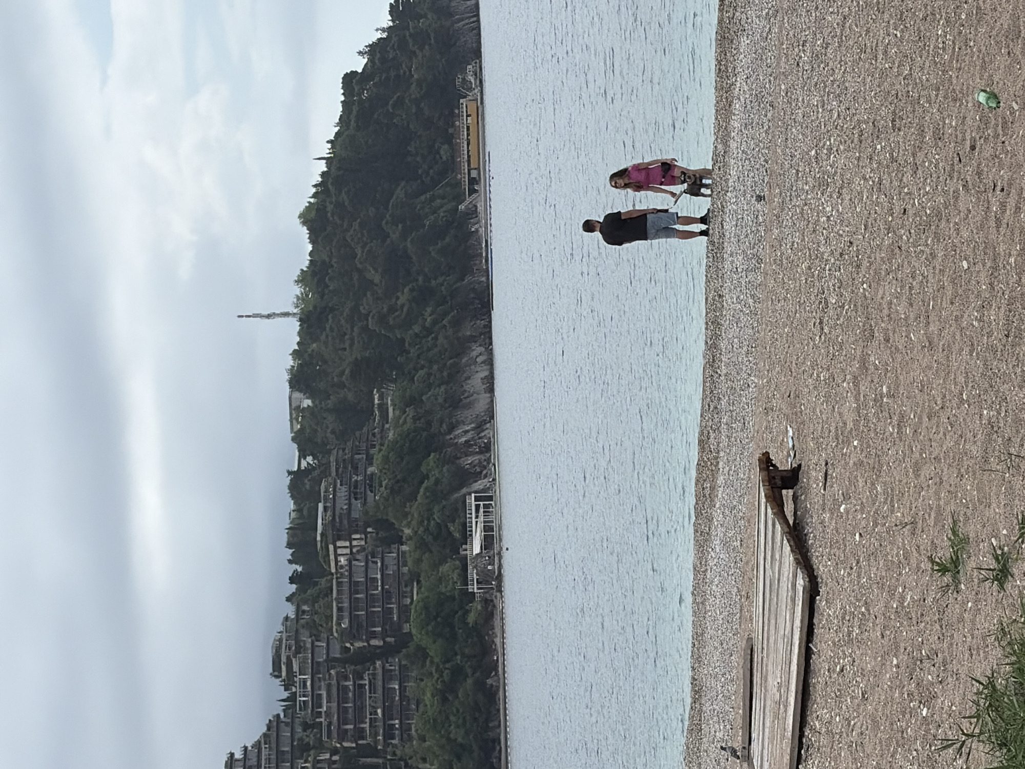The image depicts a beach scene in Budva, Montenegro. A man and a woman are standing on a pebble beach along the waterline, appearing to be looking out at the water. The man is wearing a dark shirt and shorts. The woman is wearing a pink shirt. The water is calm. In the background, a steep, wooded hillside rises sharply from the coastline. A complex of buildings is visible on the hillside. Part of a decaying wooden structure is on the beach. The sky is overcast. A communication tower can be seen on the hill.