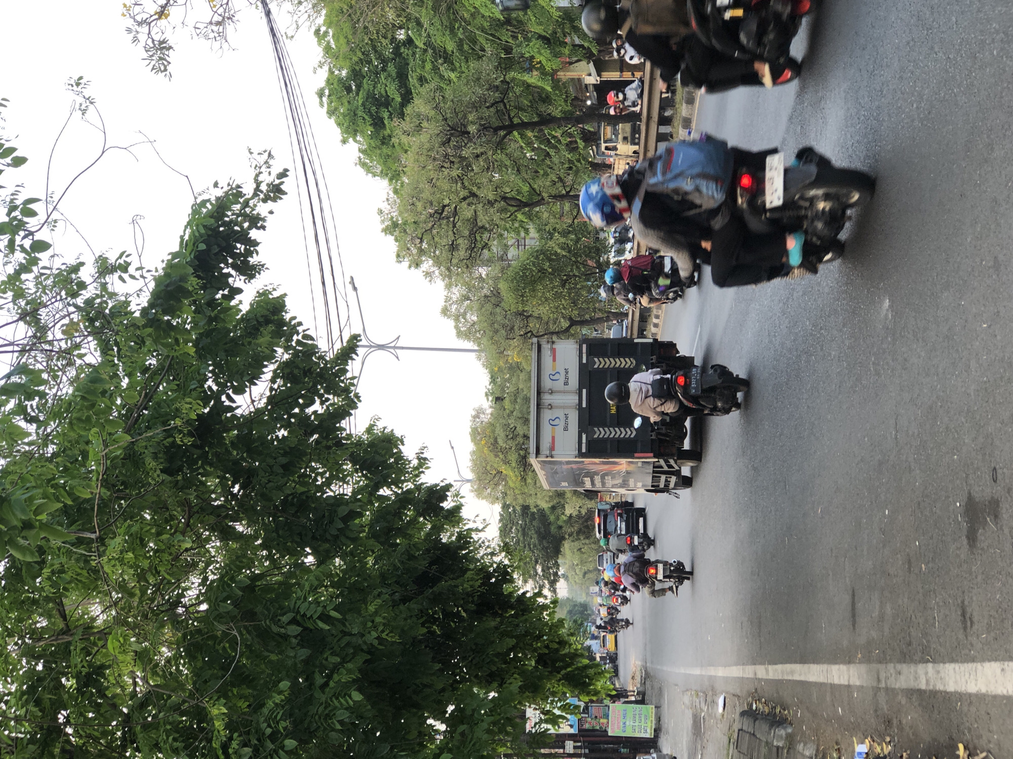 The image captures a street scene in Kludan, Indonesia, with several motorcycles and a truck on a paved road. Lush green trees line the left side of the road, their branches partially obscuring the sky. Power lines are visible overhead. The road is populated with various vehicles, including multiple motorcycles, some carrying one or two riders, and a large truck. One motorcyclist in the foreground is wearing a blue backpack. The truck has "Biznet" branding on its side. The overall atmosphere suggests moderate traffic.