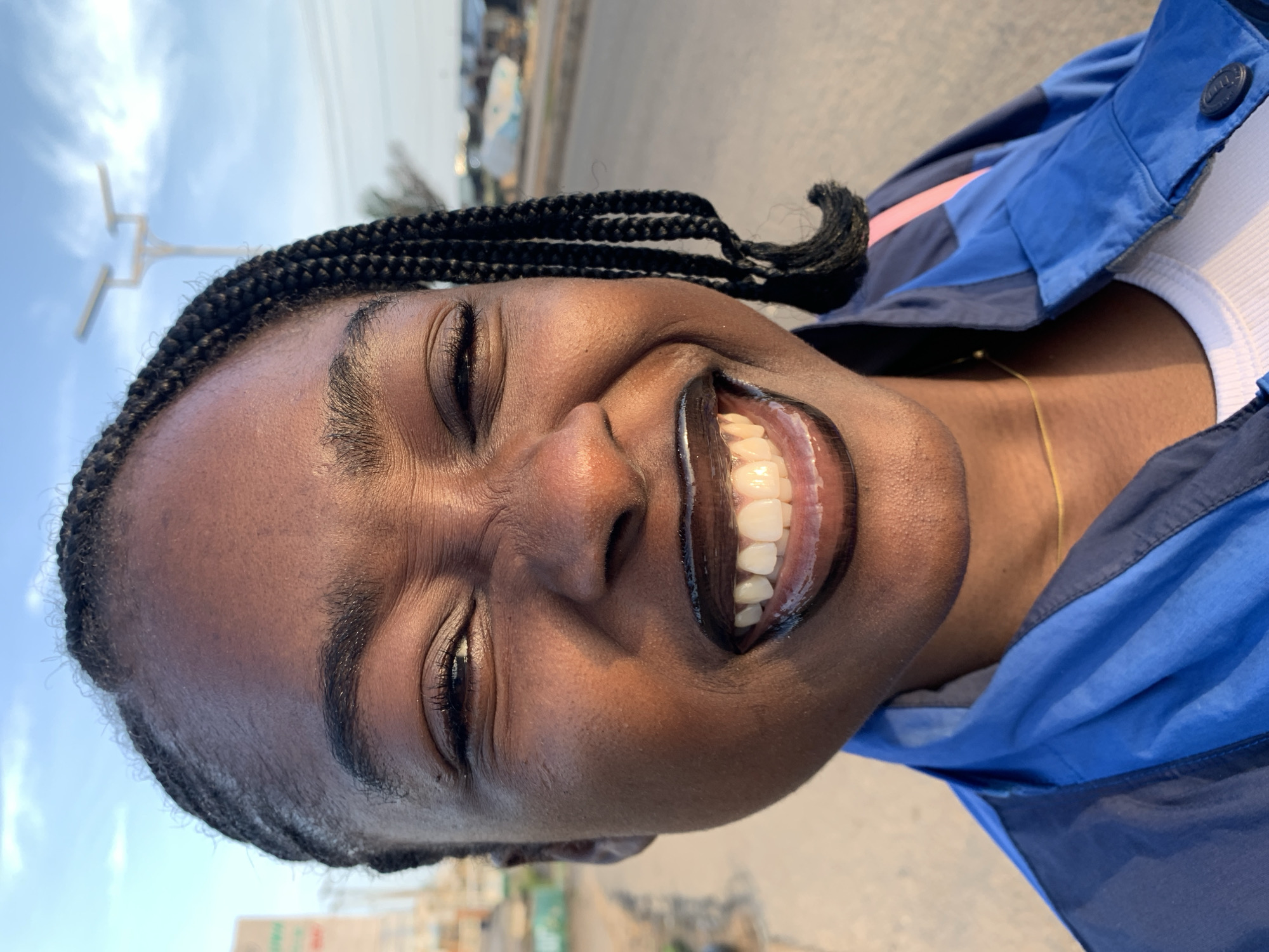 A close-up, eye-level, outdoor portrait captures a young Black woman with braided hair and dark lipstick smiling broadly. She is facing forward, and her eyes are squinted in mirth. The woman is wearing a blue jacket over a white ribbed shirt, and a thin gold chain is visible around her neck. The background, blurred due to the shallow depth of field, suggests an urban environment. In the distance, a streetlamp with two lights is visible, along with a road and buildings. This scene is set in Rafin Par, Nigeria.