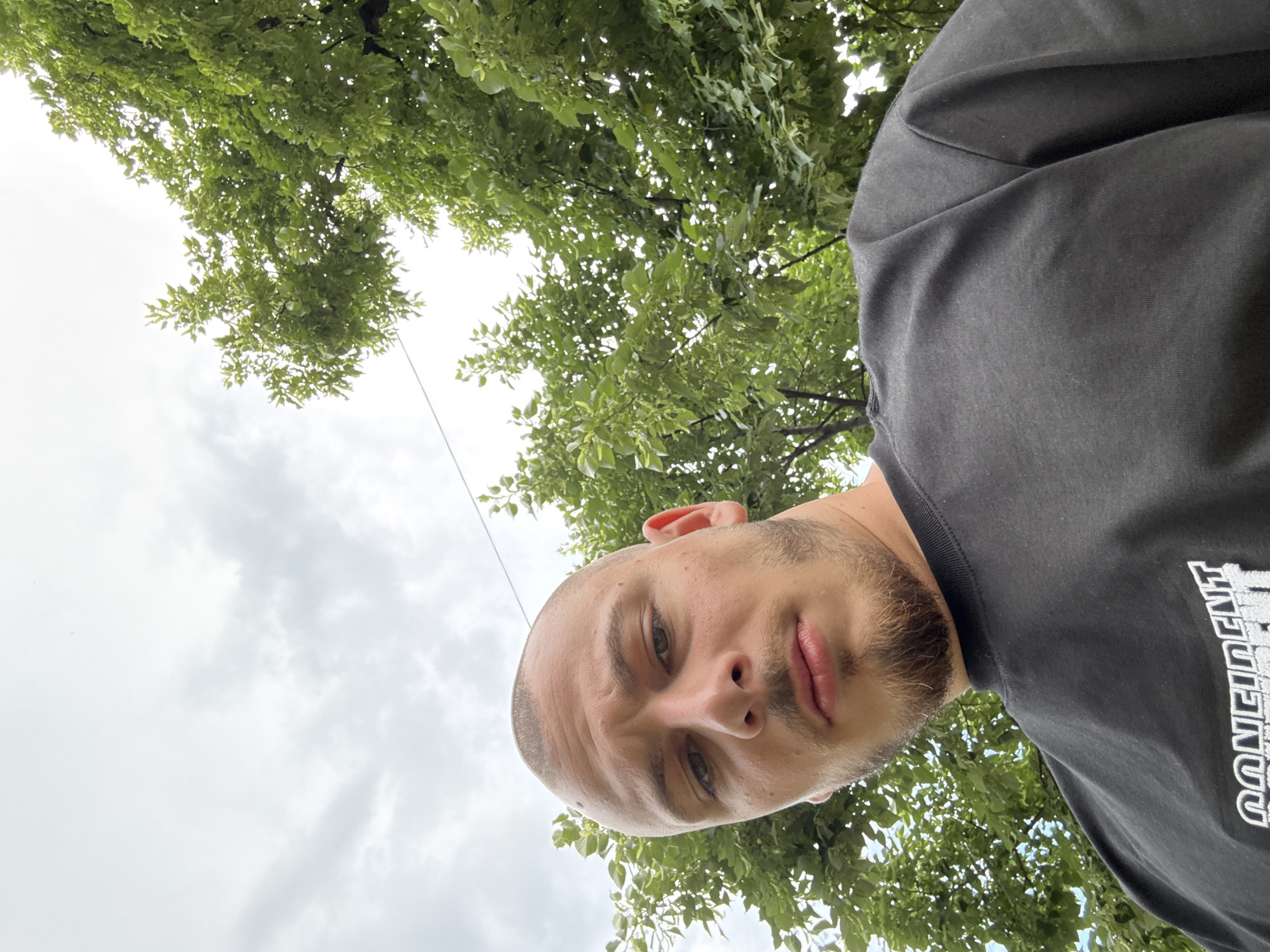 A man with a shaved head and a trimmed beard looks down at the camera from a low angle. He is wearing a black t-shirt with white lettering that reads "Confident." The background consists of green leafy trees and a cloudy sky. The image appears to have been taken outdoors. The specific location in Stara Zagora, Bulgaria, cannot be determined from the visual information provided.