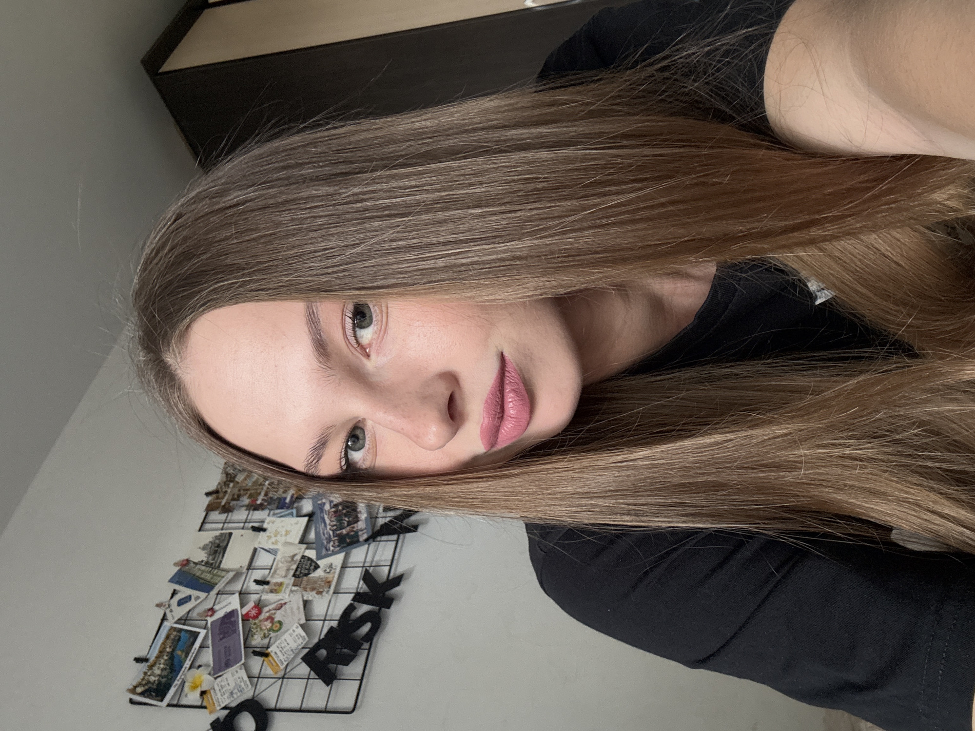 The image shows a close-up, low-angle selfie of a young woman with long, brown hair, taken indoors. She is wearing a black t-shirt and has a fair complexion. Her left eye, a green-blue color, is visible, as is the bridge of her nose and her lips, which are painted with a matte pink lipstick. Her eyebrows are well-defined and dark. Her hair is parted and falls across her face, obscuring part of her forehead and one eye.
In the background, to the left of the subject, is a black wire grid board on a light-colored wall, adorned with numerous small photos and postcards, as well as some black letter cutouts. One visible letter cutout appears to spell "RISK." The grid and photos suggest a personal display, possibly in a bedroom or living space. Above and behind the woman's head is a dark wooden surface, possibly the top of a cabinet or shelf. The lighting appears to be natural, coming from an unseen source, and the overall atmosphere is casual and relaxed. The image is vertically oriented. There is no indication of the time of day or weather conditions. The location context provided is Perm, Russia.