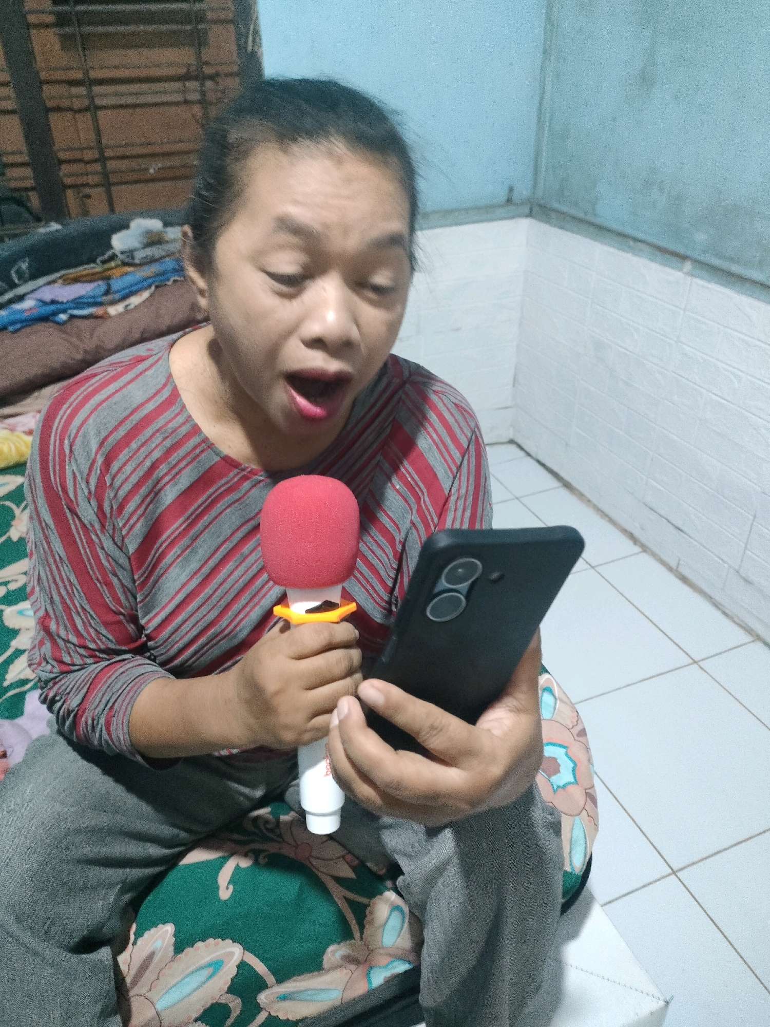 A person is sitting on a patterned cushion and holding a small red microphone in their right hand and a black smartphone in their left hand. They are wearing a gray and red striped long-sleeved shirt and gray pants. The person's mouth is open as if they are speaking or singing. The background appears to be a room with white tiled walls and a tiled floor. The lighting is dim, suggesting it might be indoors at night or in a poorly lit space. The overall impression is that of someone engaged in a personal activity like karaoke or recording. This image does not contain any identifiable landmarks or features that would allow for pinpointing a specific location within Pekanbaru, Indonesia.