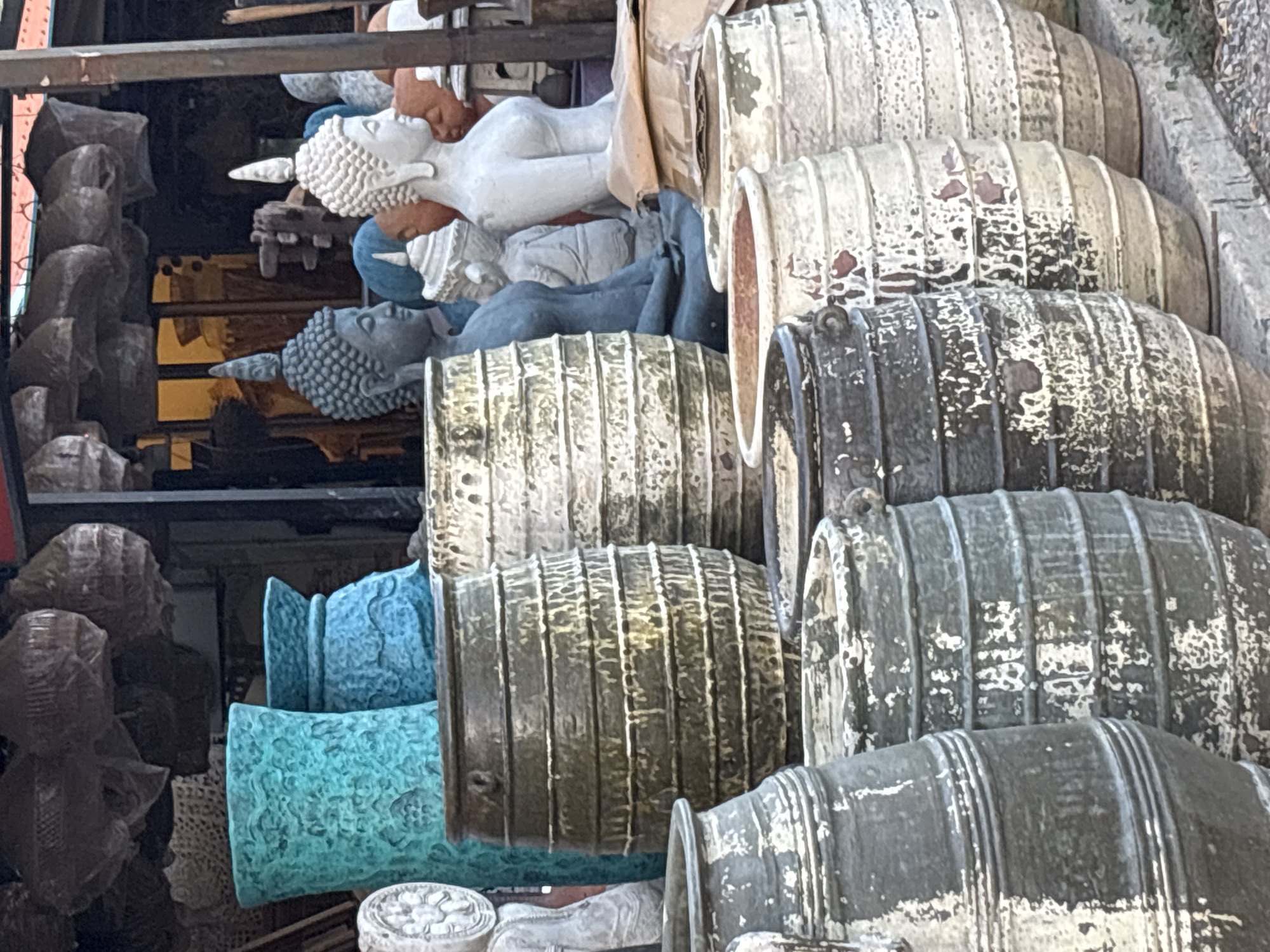 The image depicts a scene in Baan Bo Phut, Thailand, showcasing a collection of various objects and statues stacked together, apparently for sale. In the left-hand side of the image is the side-view of various dark gray stone statues, most notabley a side profile of buddha with a rounded head. Further towards the right, statues of buddha laying sideways are visible. One is white, and one is light blue. In front of the statues, stacked cylinders, likely decorative pots or vases, are positioned. These items have ridged exteriors and worn-down finishes with visible signs of aging. The general atmosphere suggests an outdoor marketplace or a vendor's stall.