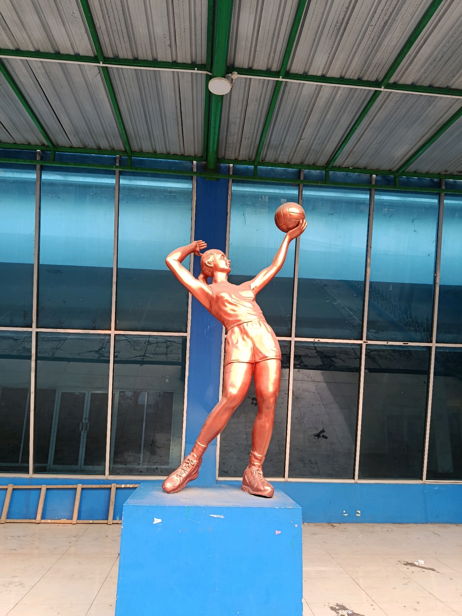 A full-body bronze statue of a female volleyball player mid-serve is centrally located on a blue pedestal. The statue, with a metallic copper finish, depicts a woman in athletic wear, holding a volleyball above her head with her right arm extended. Her left arm is bent behind her head, and her body is arched in motion. She wears shorts and athletic shoes. The statue stands in front of a building with large, blue-tinted glass windows framed by white mullions. The windows reflect the sky and the interior of the building, which appears to be an indoor sports facility or covered public space. Above the windows, a silver corrugated metal roof is supported by green metal beams, with a white pipe and a circular light fixture visible. To the left of the statue and below the window line, a wooden stepladder rests against the blue wall. The ground is tiled with square beige and white tiles, with some scattered debris. The scene suggests an outdoor or semi-outdoor area of a sports complex. This statue is located in front of the Graha Cakrawala building at the State University of Malang (UM) in Malang, Indonesia.