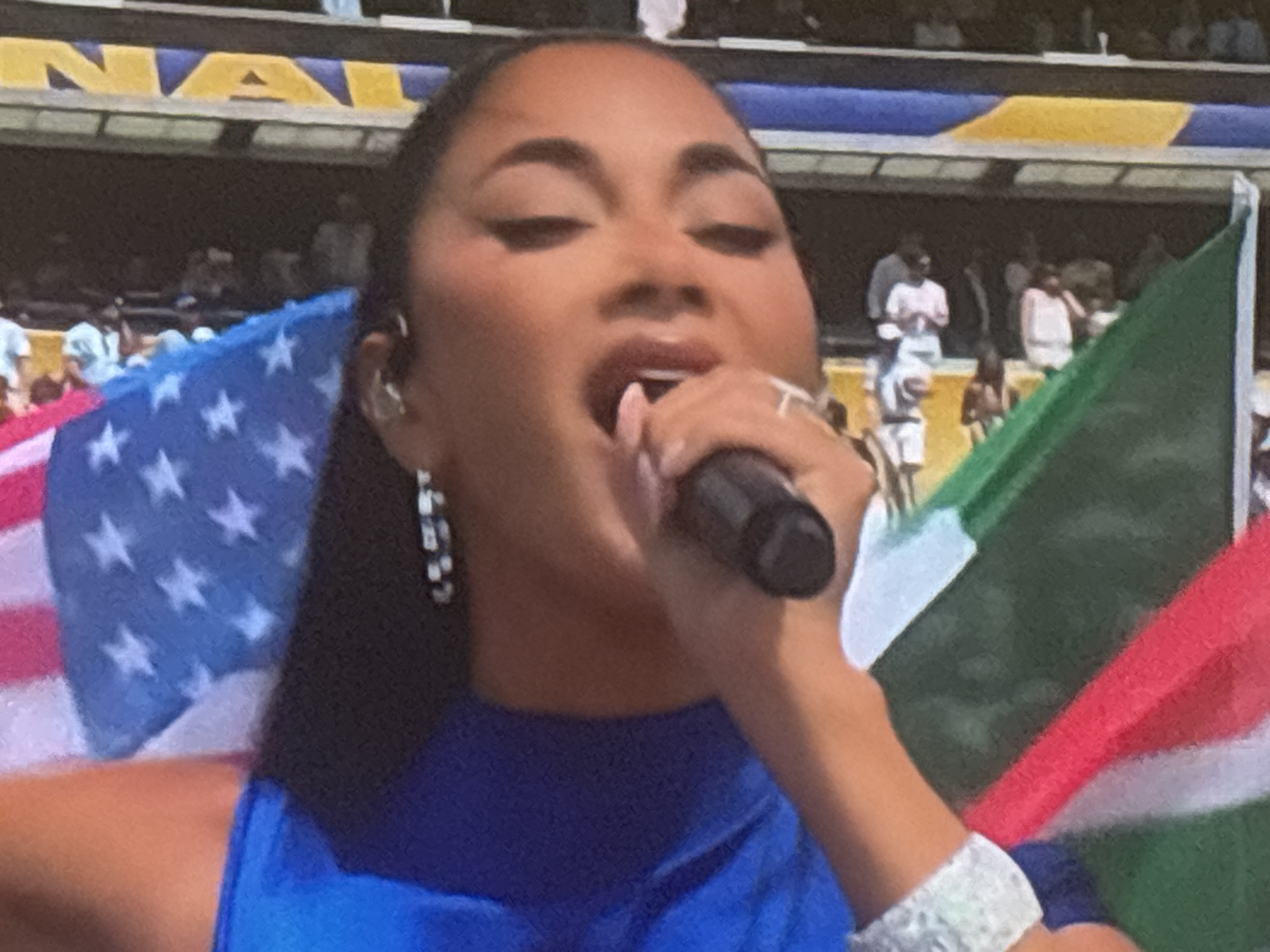 The image shows a close-up of a woman singing into a microphone. She is wearing a blue top and has several pieces of jewelry on, including earrings and bracelets. Behind her, flags of the United States and South Africa are visible, as well as a stadium crowd. The woman appears to be performing on a stage during an outdoor event. The location in Burgas, Bulgaria, is not ascertainable from the image.