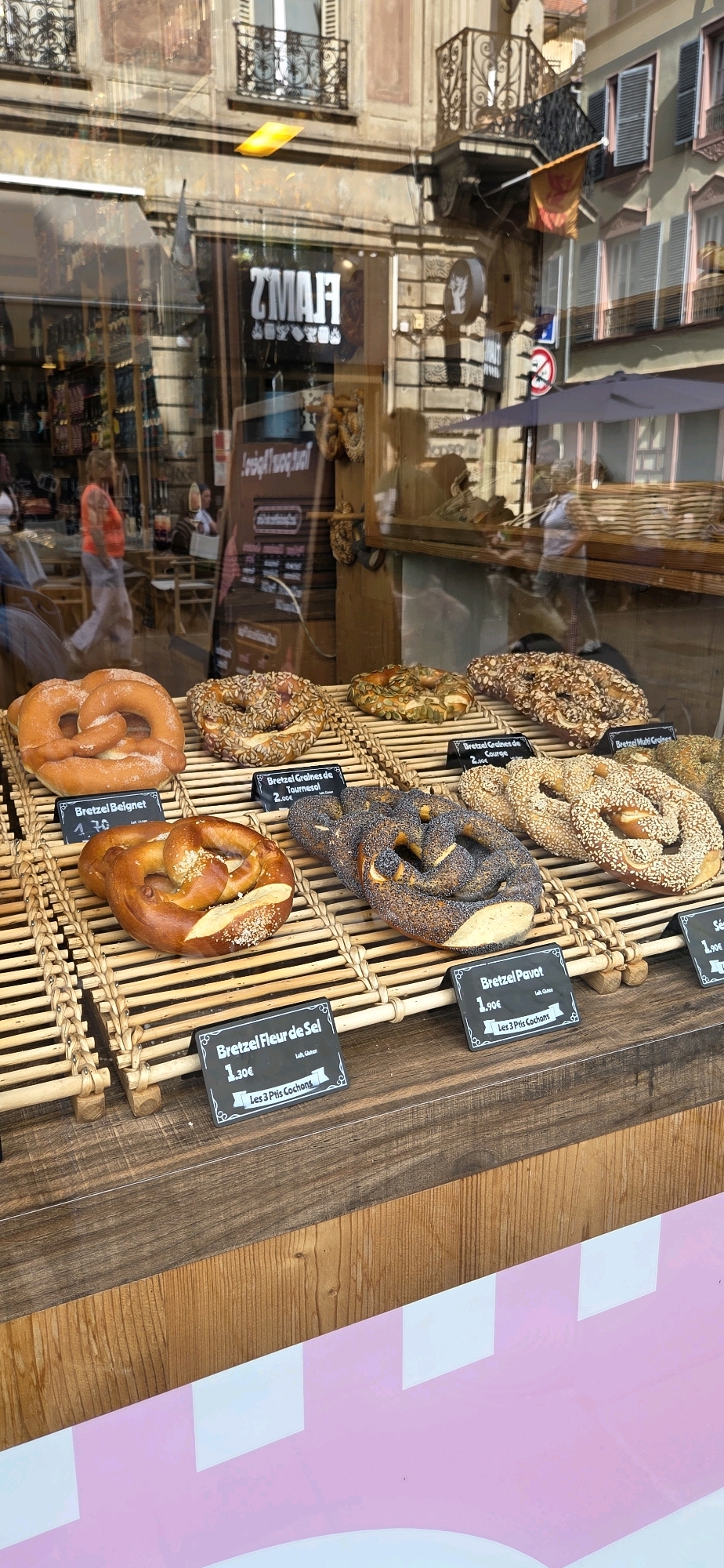 The image shows a close-up view of a bakery window display in Strasbourg, France. Various types of pretzels are arranged on tiered wooden shelves. Some pretzels are plain, while others are topped with seeds like sunflower seeds, poppy seeds, and pumpkin seeds. Price tags are visible for some of the pretzels, indicating prices in Euros. Behind the display, the interior of the bakery is reflected in the glass, showing shelves with more baked goods and possibly merchandise. In the background, seen through the window, are reflections of buildings with typical Alsatian architecture, including ornate balconies and shuttered windows, suggesting a street in Strasbourg. People are visible in the reflection walking inside the bakery and on the street outside. The overall scene is well-lit, suggesting daylight.