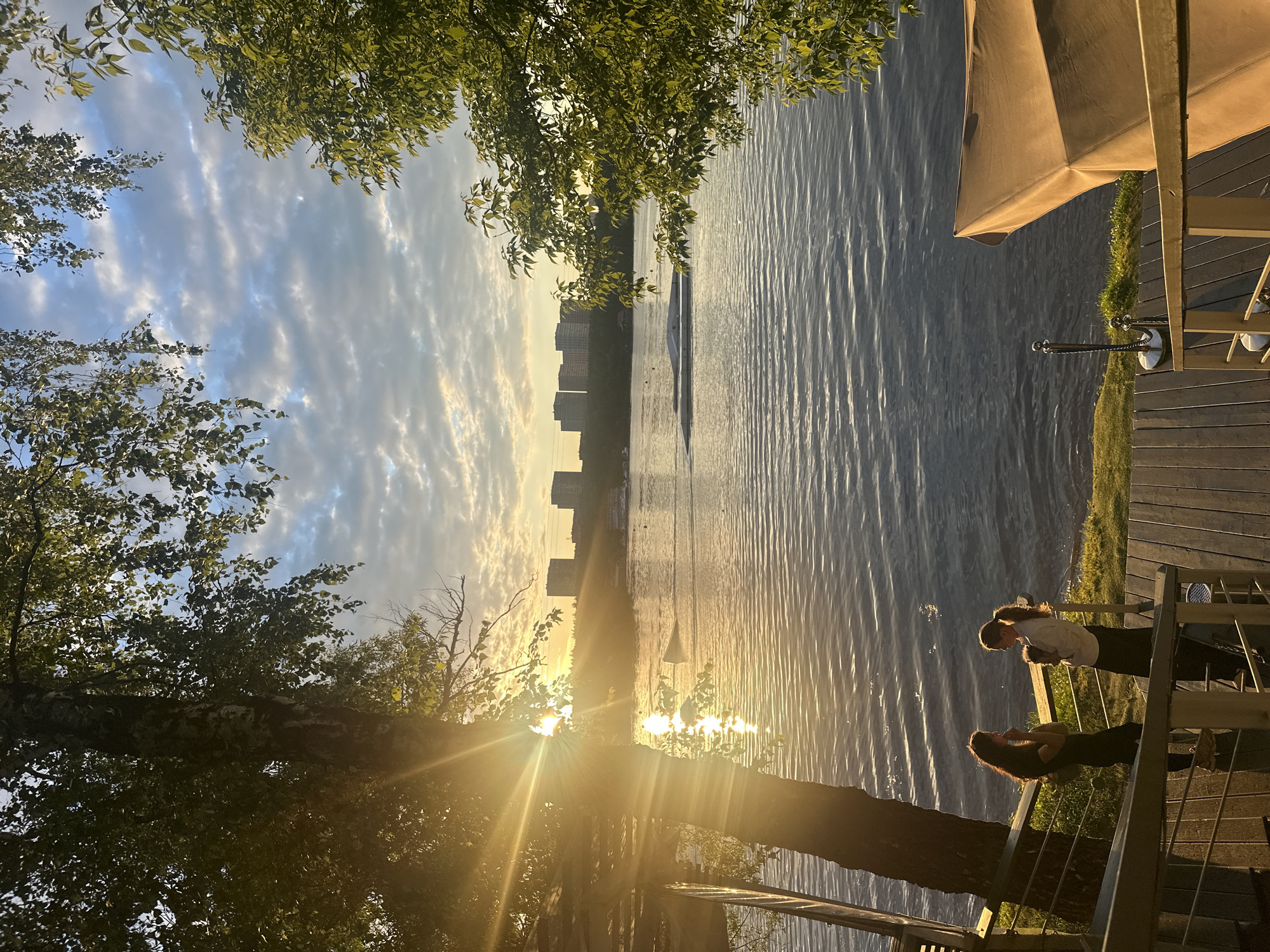 The image captures a sunset scene by a body of water, likely in Moscow, Russia. The sun is low in the sky, casting a warm golden glow across the water and creating lens flares through the surrounding trees. The sky is a mix of blue with scattered, wispy clouds, some of which are illuminated by the setting sun.
In the distance, a city skyline with several tall buildings is visible across the water. The water itself is calm with gentle ripples, reflecting the light. A small white structure, possibly a boat or a floating platform, is visible in the water.
In the foreground, on a wooden deck or balcony, two young women are standing and facing each other. They appear to be engaged in a conversation, with one woman leaning forward and the other looking up. The deck is furnished with railings and a large umbrella, suggesting it is part of an outdoor seating area, perhaps a cafe or restaurant. The overall mood is peaceful and serene, enhanced by the soft lighting of the sunset. There is no visible text in the image.