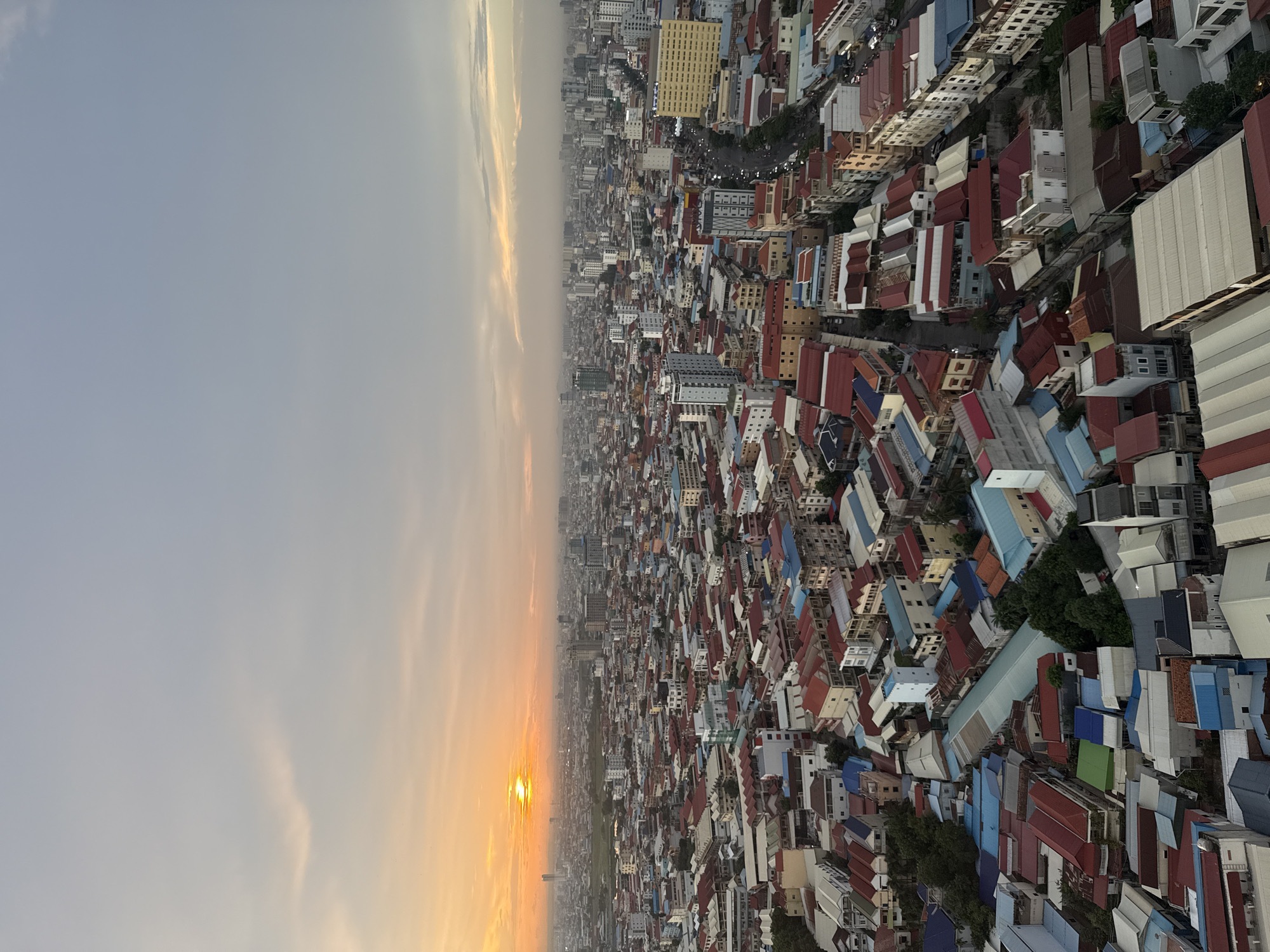 This is an aerial, high-angle photograph of a densely populated urban area, likely Khan Mean Chey, Cambodia, during sunset. The sky is filled with soft, muted colors of orange, yellow, and grey from the setting sun, with a few wispy clouds. The cityscape is dominated by a vast expanse of buildings with a variety of roof colors, including red, blue, white, and grey. Many of the structures appear to be low-rise residential or commercial buildings, packed closely together. In the distance, taller buildings and skyscrapers punctuate the horizon, suggesting a mix of older and newer developments within the city. A road with visible vehicle traffic winds through the buildings in the mid-ground. The overall impression is of a bustling city at dusk, with a warm, hazy glow from the setting sun.