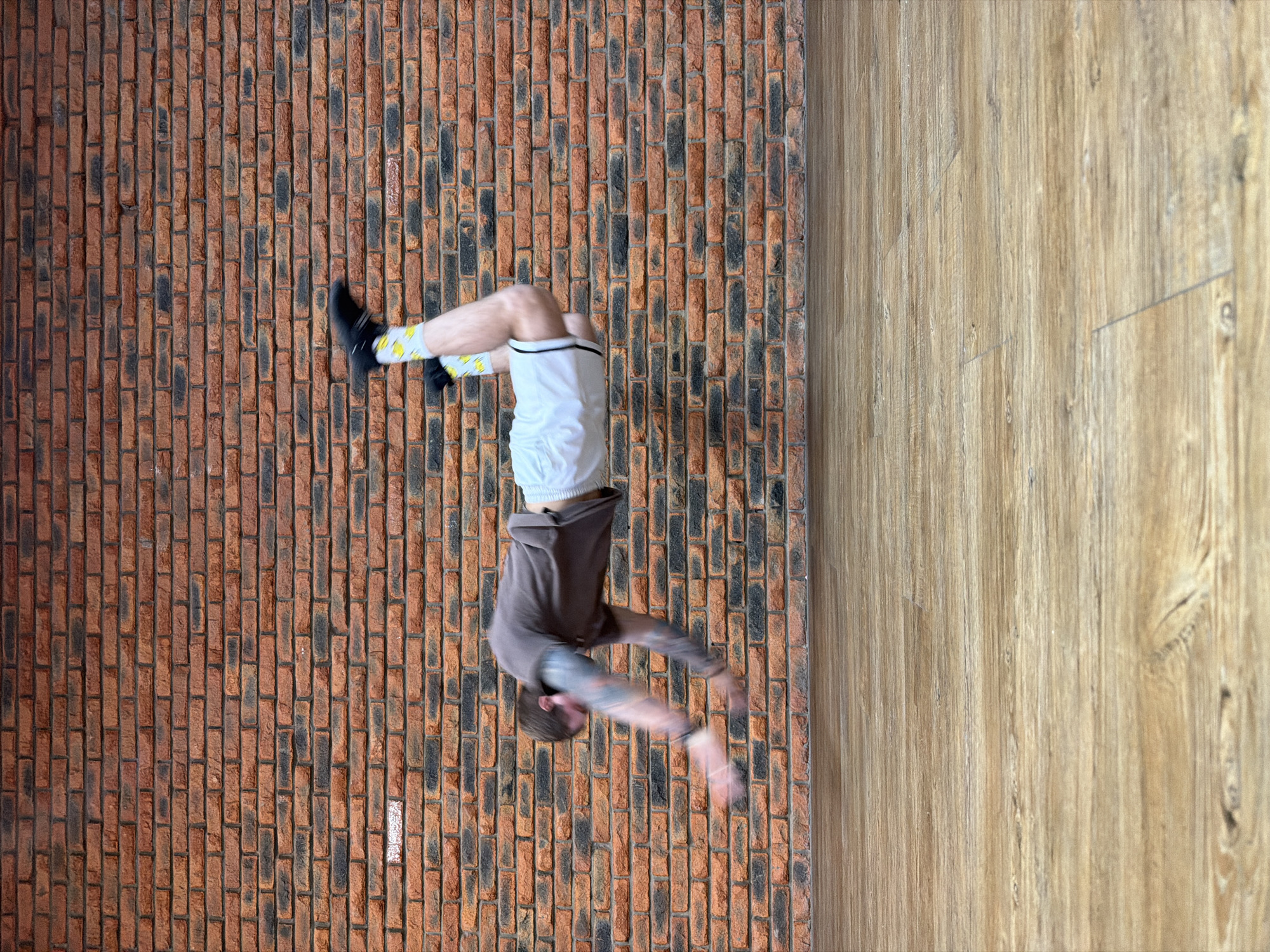 The image depicts a man in motion, captured mid-air in what appears to be a backflip. He is wearing a brown hoodie, white shorts, and socks with a design. His arms are extended, and the motion is blurred, suggesting rapid movement.
The location features a brick wall on the left side and a light wood-patterned surface on the right. The image is rotated, giving the impression that the person is performing the flip on a vertical surface.
There are no specific details indicating the city of Chaikovsky, Russia, in the image.
