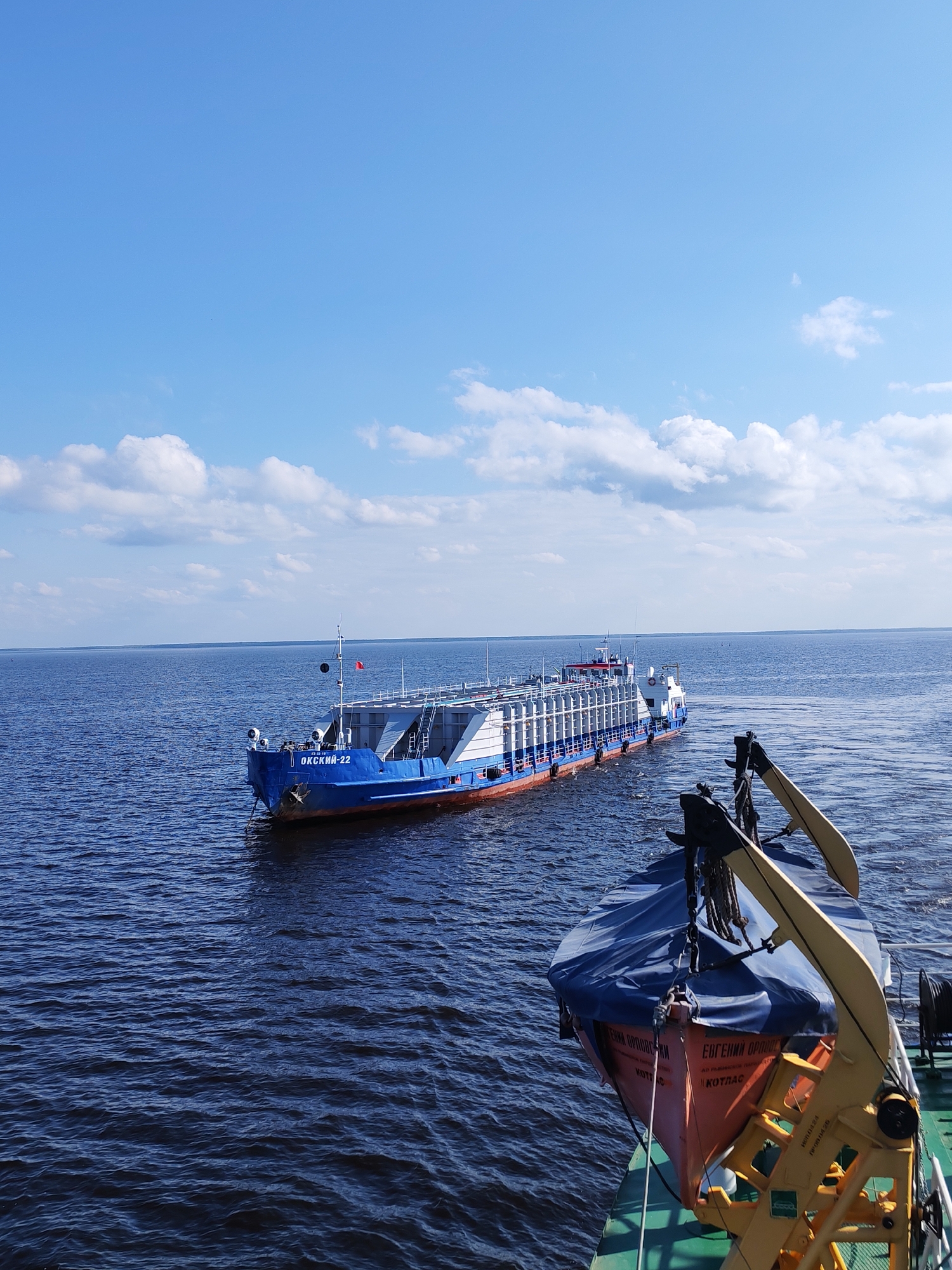 The image depicts a water scene with two vessels on a body of water, likely a river or lake. The sky is blue with scattered white clouds. The vessel in the foreground appears to have a covered lifeboat with lifting apparatus. The name “Евгений Орловски” is partially visible on the lifeboat. The vessel further away is a blue and white barge-like structure with the name "Окский-22" visible on the bow. The water is dark blue and slightly choppy. The image was taken in Bol'shie Mkhi, Russia.