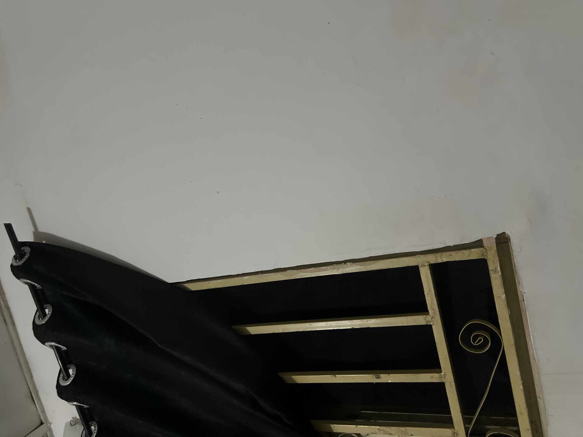 A black curtain with metal grommets is draped across the top left of the frame, obscuring a barred window. The window bars are painted a dull yellow and are arranged vertically with a horizontal bar across the bottom. A decorative scrollwork element is visible on the lower horizontal bar. The wall surrounding the window is painted a light, neutral color and appears somewhat textured. The scene is dimly lit, suggesting it is nighttime or indoors with limited light. The overall impression is one of security or privacy due to the bars and curtain.