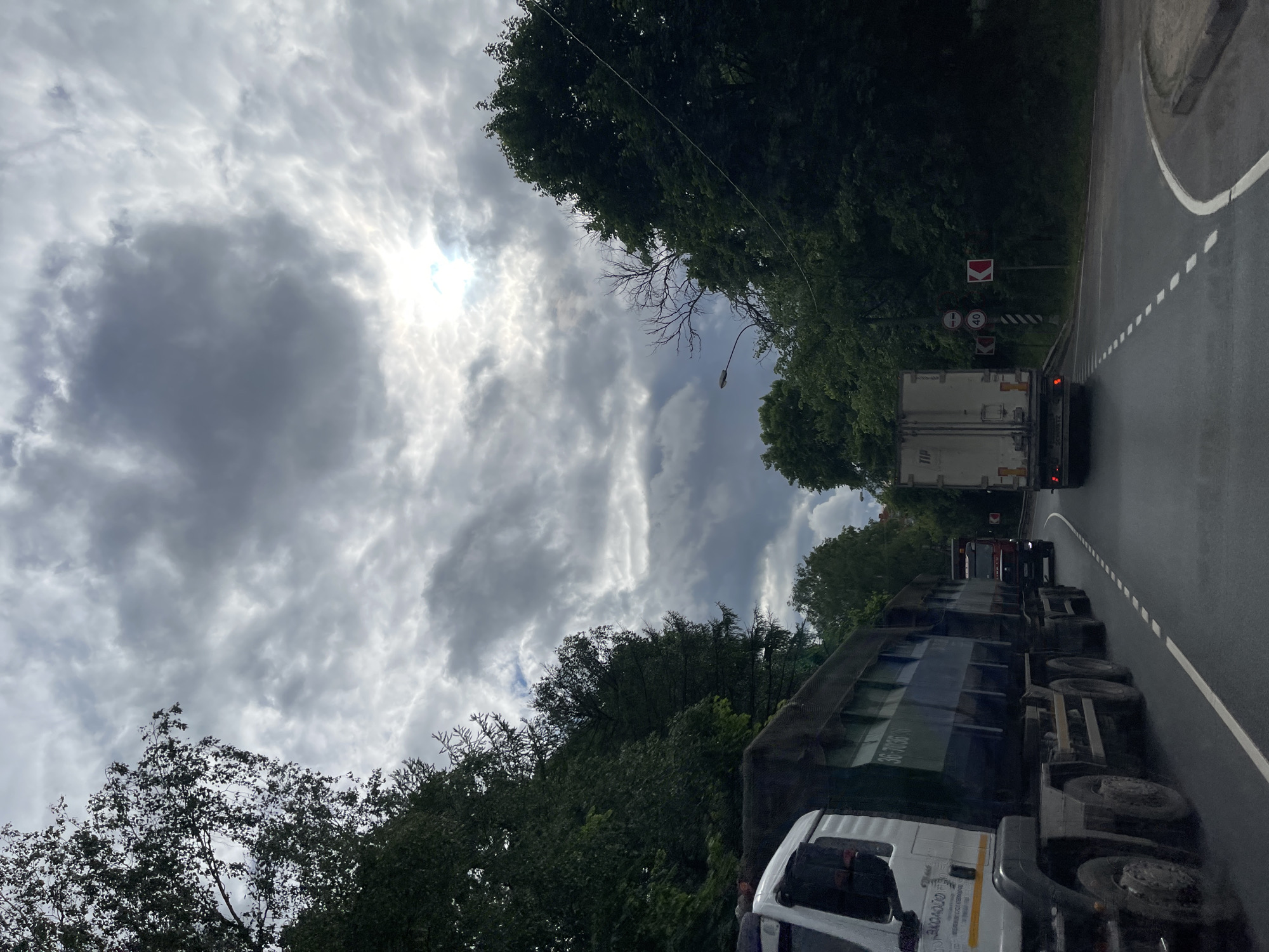 The image shows a road in Semyonovskoye, Russia, under a cloudy sky. Several trucks are on the road, including a white one in the foreground. Trees line both sides of the road. There are traffic signs visible near a curve in the road further ahead. The sun shines through the clouds in the sky.