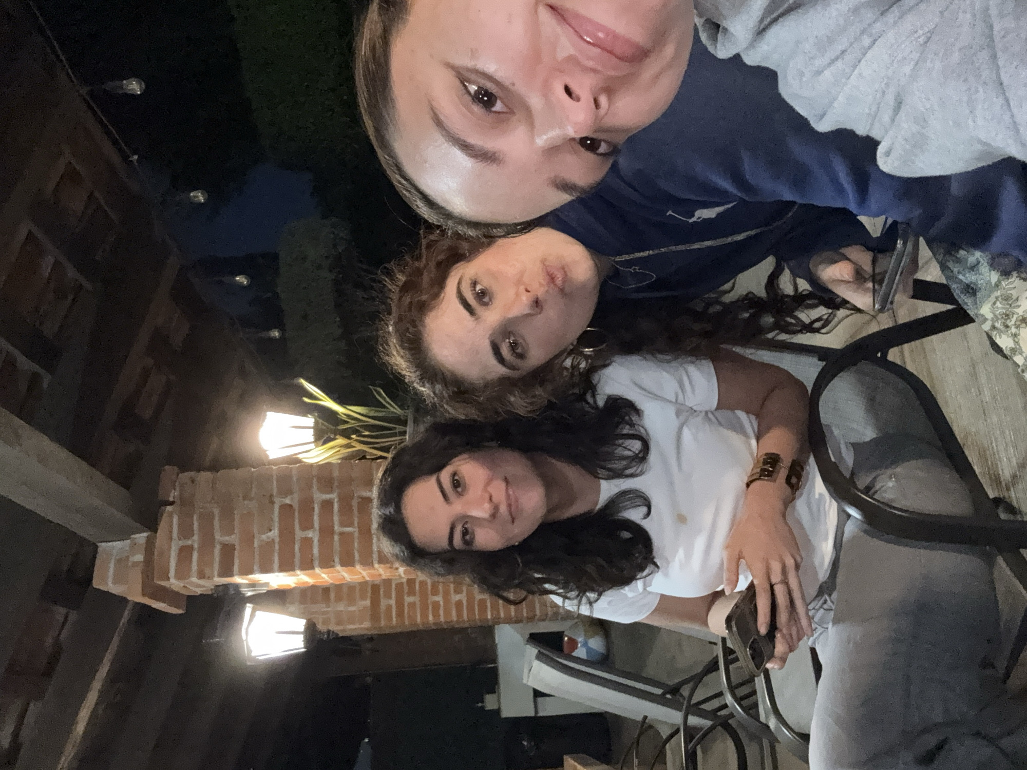 Three women pose for a selfie. The woman in the foreground wears a white t-shirt and jeans and has long dark curly hair. The woman in the middle has long curly brown hair. The woman in the background wears a grey sweatshirt and a blue shirt. The photo was taken outdoors at night in Puebla, Mexico. A brick structure is visible behind the women. Lights illuminate the scene.