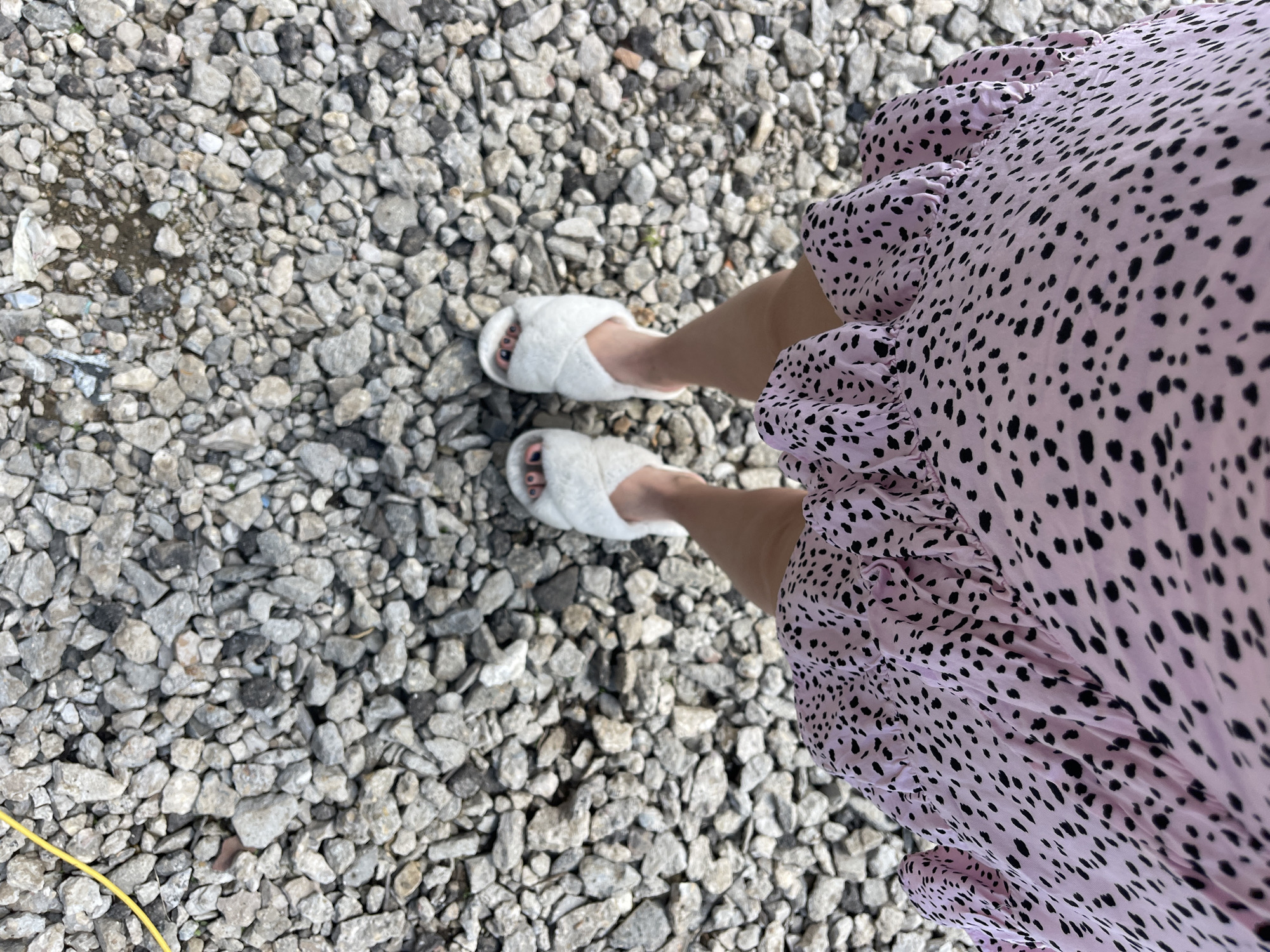 A top-down shot shows a person standing on a gravel surface, likely outdoors. The person is wearing white, fuzzy slippers. The person is also wearing a light pink dress with black polka dots, which are predominantly visible on the right side of the frame. A yellow wire is visible on the left side of the frame near the gravel. The location is Мосметаллоконструкция Онт, Russia.