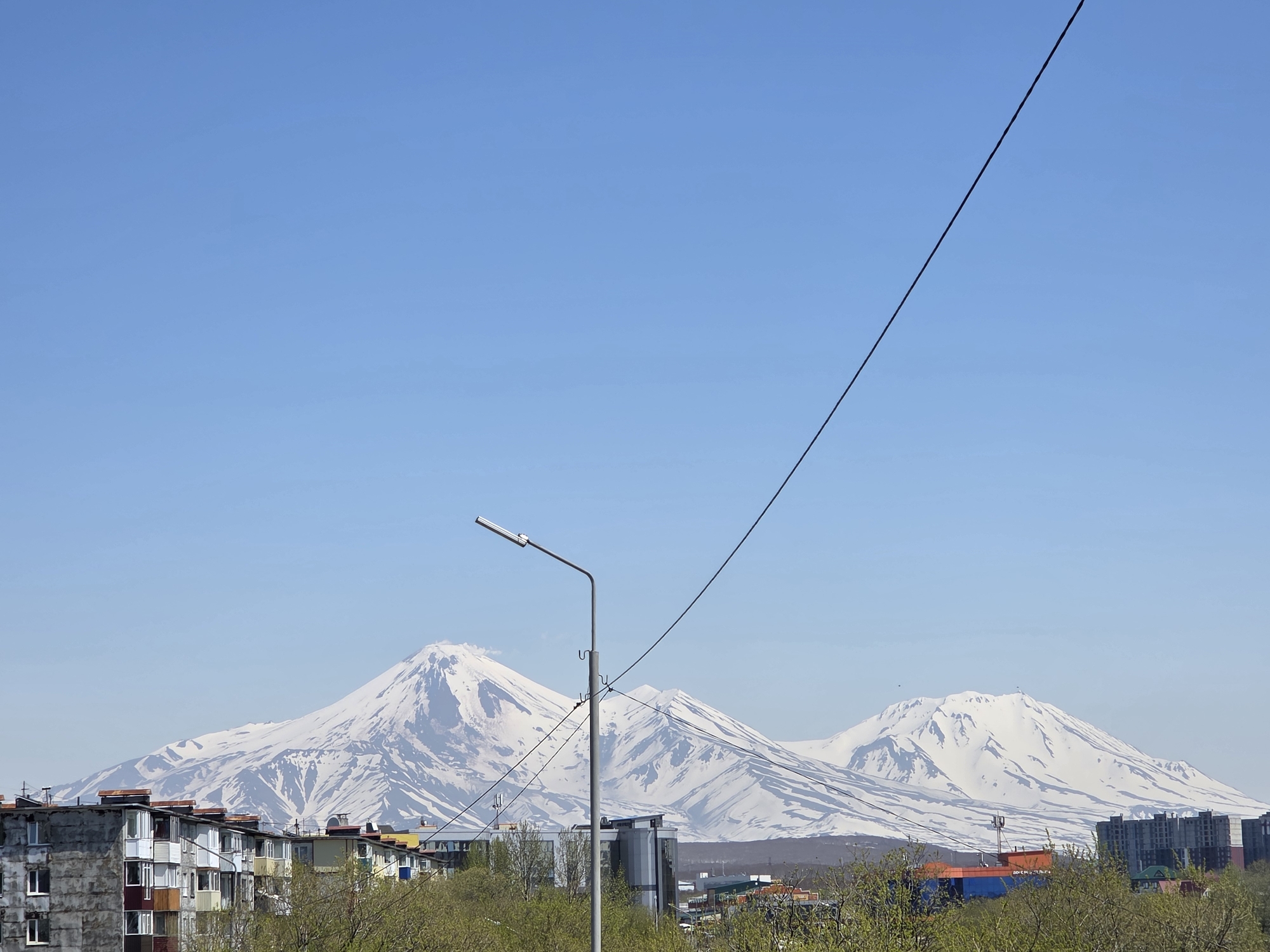 The image displays a clear, bright day in an urban environment set against a dramatic natural backdrop. In the background, three prominent, snow-covered volcanic peaks dominate the horizon under a cloudless, light blue sky. The snow cover on the mountains appears patchy, revealing darker rock beneath.
In the mid-ground, a gray metal street light pole with a horizontal luminaire pointing left stands centrally. Multiple dark utility wires extend from and around this pole, with one thick wire stretching diagonally from the pole towards the upper right corner of the image. Below the mountains and behind the street light, various buildings with industrial or commercial appearances are visible, including one distinct structure with orange and blue sections. Green trees and foliage are scattered across the mid-ground, suggesting a transition from urban to natural landscape.
The foreground, particularly the lower left quadrant, features a row of multi-story residential apartment buildings. These structures exhibit varied facade colors and numerous balconies, some enclosed and others open, indicative of functional urban architecture. No discernible people, animals, or active vehicles are visible within the frame. The location is in the city of Unknown, Unknown.