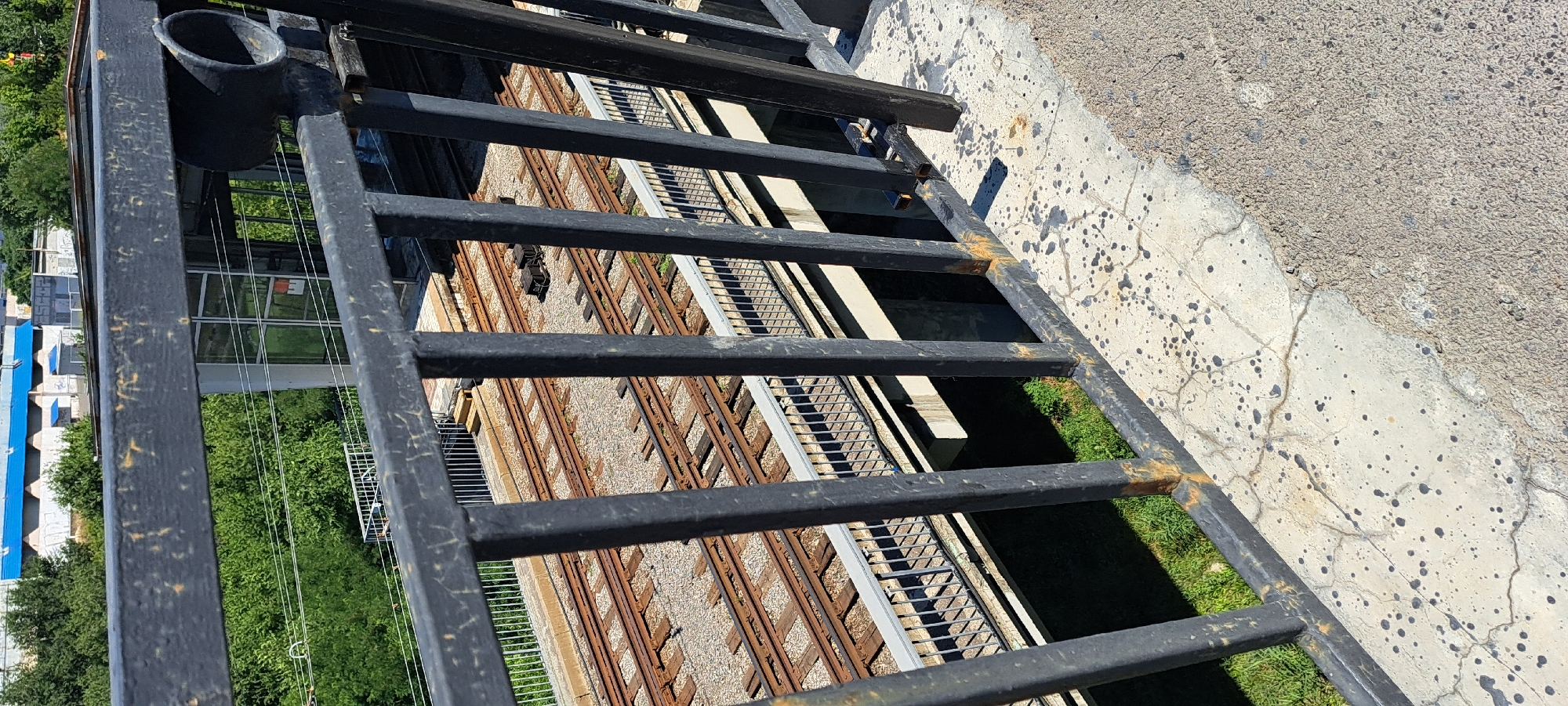 The image shows a view through a black metal fence, overlooking a set of train tracks. The fence is in the foreground, with vertical bars and a horizontal top rail. The tracks are below and slightly to the left, with several sets of parallel rails running across the frame. Some ballast stones are visible between the ties. A concrete bridge or overpass structure supports the tracks. In the background, there are trees and buildings, including a building with a blue roof and white siding. The ground in the foreground is a concrete surface with dark splatters and some cracks. The scene appears to be outdoors, during the daytime, with sunlight casting shadows.