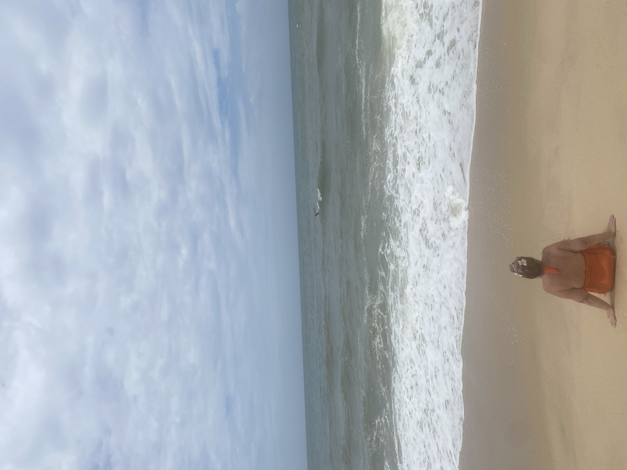 The image shows a woman sitting on a sandy beach in Mũi Né, Vietnam. She is facing away from the camera, towards the ocean. She is wearing an orange sarong and a floral hair accessory. The ocean is visible in the background, with waves breaking on the shore. The sky is cloudy. In the distance, a person is visible in the water, possibly swimming or surfing. The overall scene is a tranquil beach setting.