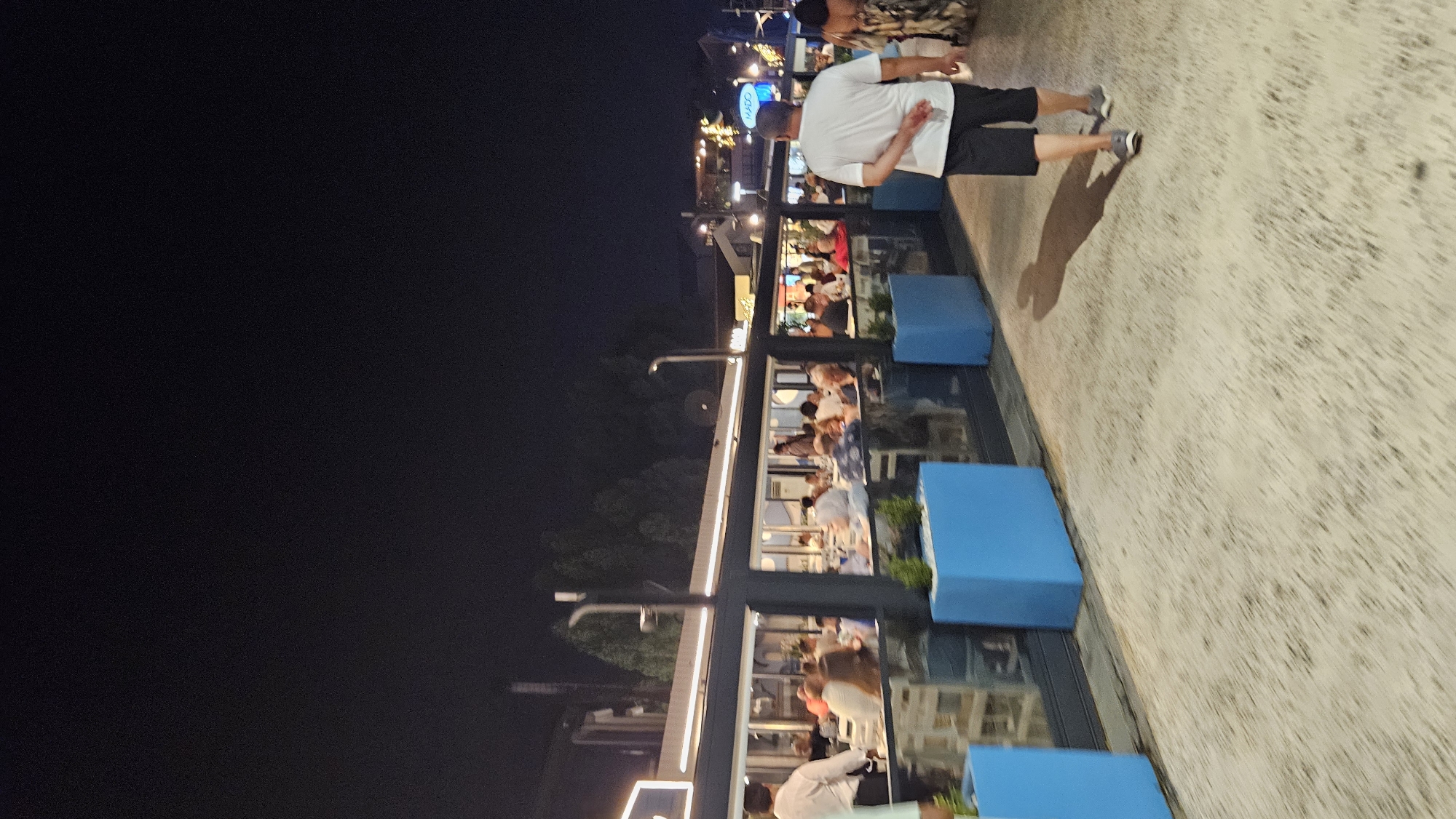 The image depicts a nighttime scene outside a restaurant in Fethiye, Turkey. A man in a white t-shirt and dark shorts is walking away from the camera on a paved walkway. To his left is a long, glass-enclosed restaurant with many patrons seated at tables. The restaurant is illuminated by interior lighting and string lights are visible in the background. Blue planters with small green plants are placed along the front of the restaurant. The sky is dark and clear.