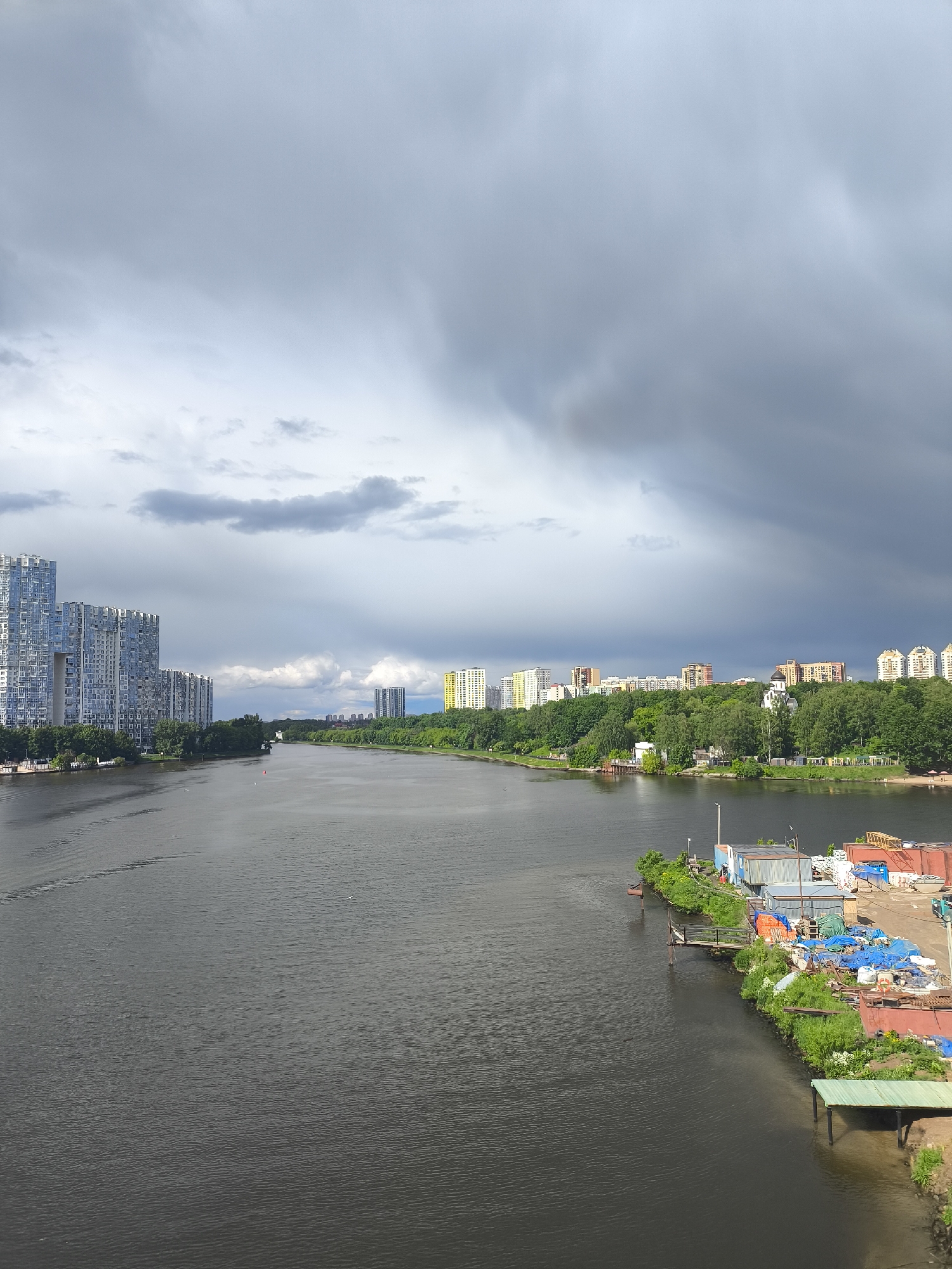 The image depicts a wide river flowing through a city, under a sky dominated by grey, ominous storm clouds. On the left bank of the river, there are tall, modern apartment buildings, with dense green trees separating them from the river. On the right bank, there is a disorganized industrial area filled with various materials covered by blue tarps and several temporary structures and docks. In the distance, there is a line of medium-rise apartment buildings set atop a hill covered with trees. The river itself is dark and still, reflecting the grey sky. The image suggests a contrast between residential areas and industrial zones along the river's edge. The city's name is unknown.