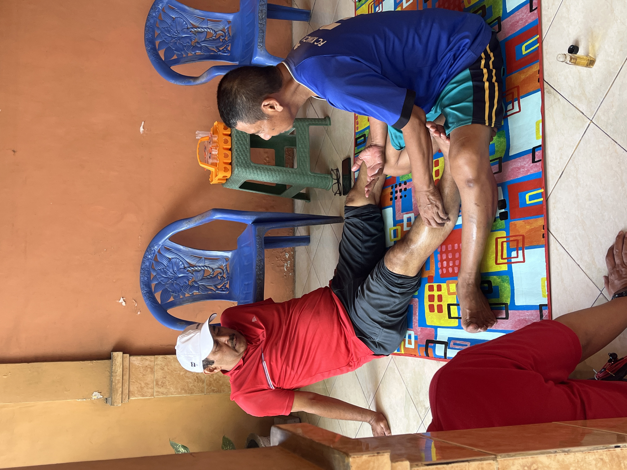 In Sidorukun, Indonesia, two men are on a colorful mat on the floor. One man, wearing a red polo shirt and dark shorts, is seated with his legs extended. A second man, wearing a blue t-shirt with the inscription "FC" visible and green and yellow striped shorts, is seated facing the first man and is massaging his leg. Behind them, two blue plastic chairs are visible against an orange wall, and a green plastic stand holds a yellow tray with small cups. A small glass bottle of oil sits on the floor in the foreground.