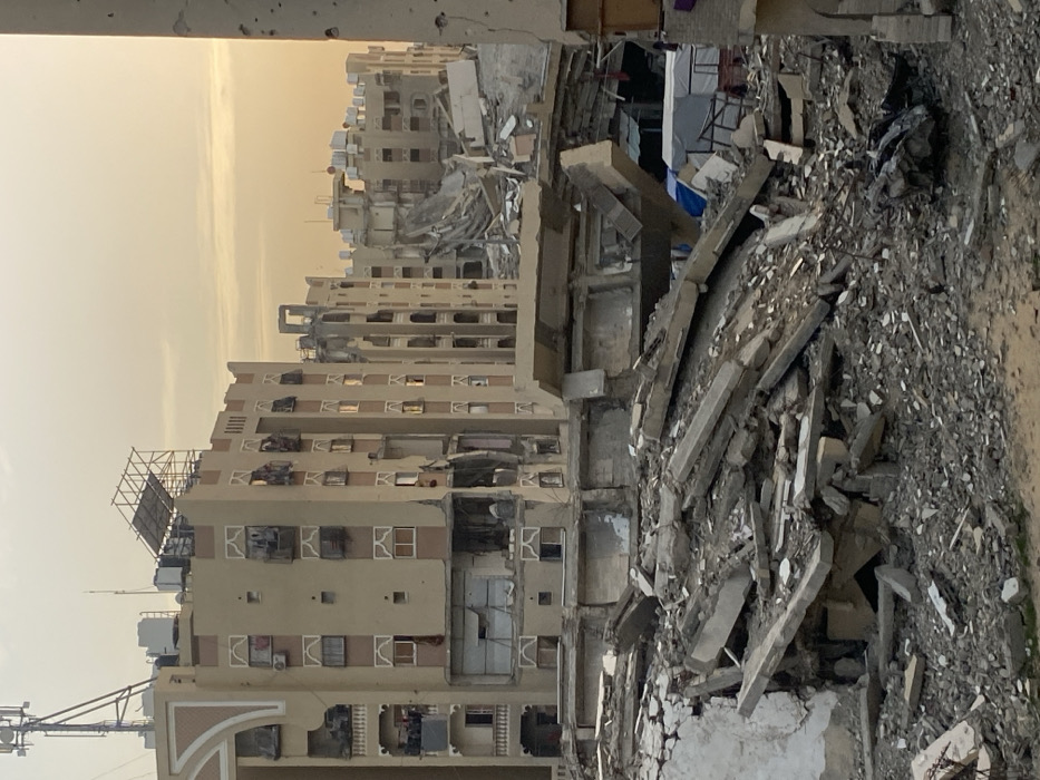 A scene of devastation. Buildings stand scarred with missing facades and shattered windows. Debris of concrete and rubble fills the foreground, evidence of destruction. The sky is hazy and muted, contributing to a somber atmosphere. The scene depicts the aftermath of conflict or disaster.