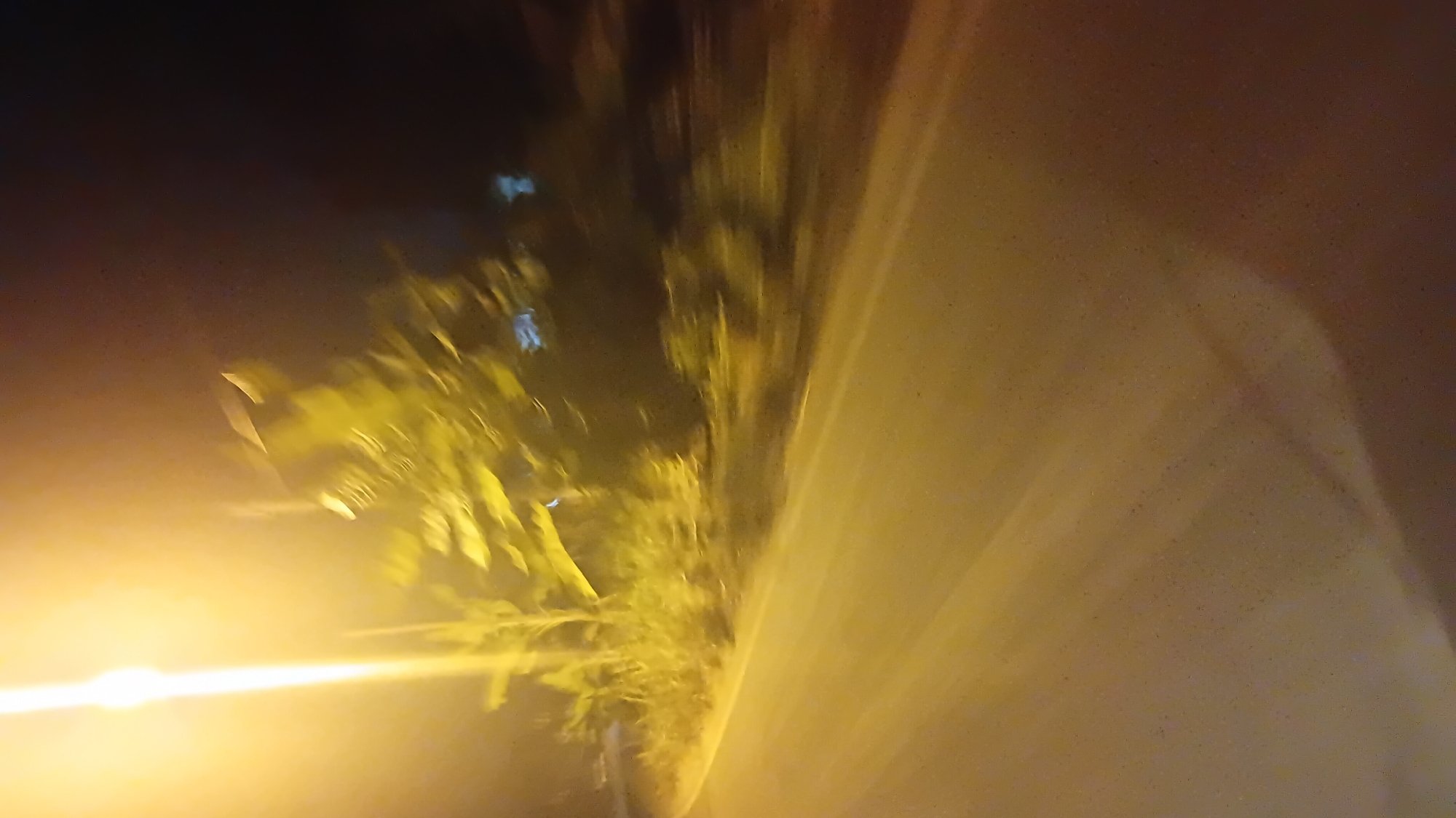 The image depicts a blurry nighttime scene of a road in Prabumulih, Indonesia, illuminated by a bright street light. The road appears to be paved and is bordered by vegetation on the left. The motion blur suggests the photo was taken from a moving vehicle. The overall impression is one of a dimly lit, possibly rural or suburban, roadside environment at night. There are no discernible people or specific landmarks.