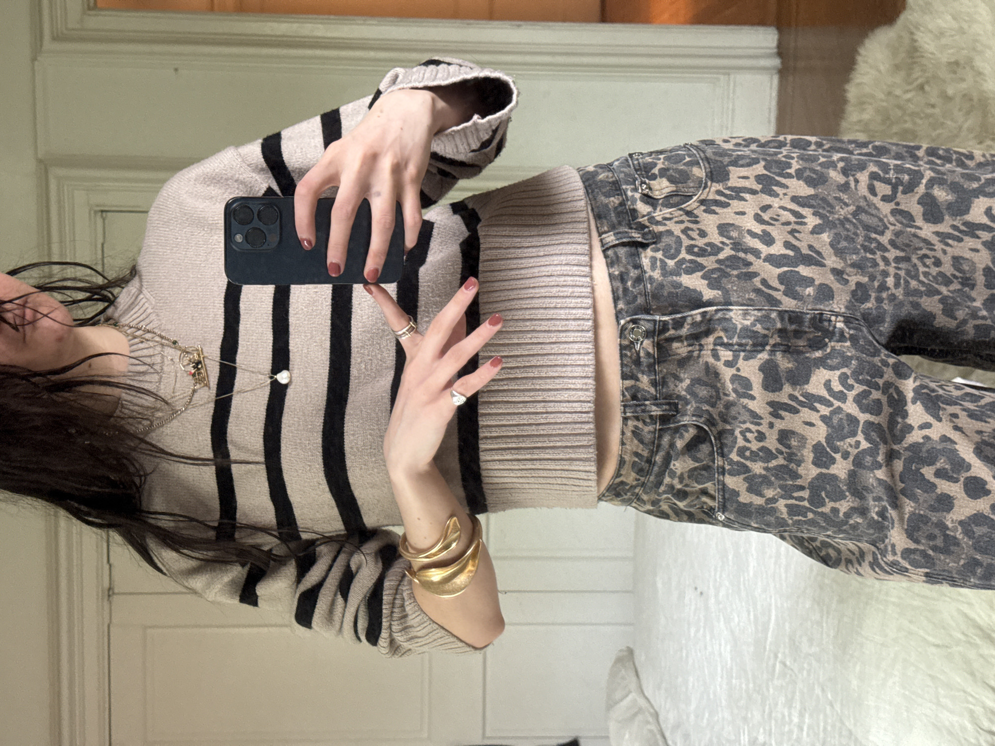 A person is captured in a mirror selfie, standing indoors. The image shows their torso, arms, and part of their head and long dark hair. They are holding a dark-colored smartphone with three visible camera lenses in their left hand, which partially obscures their face. Their fingernails are painted a dark red or burgundy color.
The person is wearing a cozy-looking, light beige knit sweater with prominent black horizontal stripes. The sweater is cropped, revealing a small section of their midriff. Below the sweater, they are wearing jeans with a distinct dark brown and black leopard print pattern.
Accessories include a large, sculptural gold-toned cuff bracelet on their right wrist, and several rings on their fingers, including a silver-toned ring on the left ring finger. A delicate silver-toned necklace with small pendants, one appearing to be a white pearl or bead, is visible around their neck.
The background, reflected in the mirror, suggests an indoor setting with light-colored walls, possibly featuring a white door or door frame on the left. A fluffy, light-colored rug or fabric is visible on the floor to the right. The overall impression is a casual, fashion-focused self-portrait. No text is visible in the image.