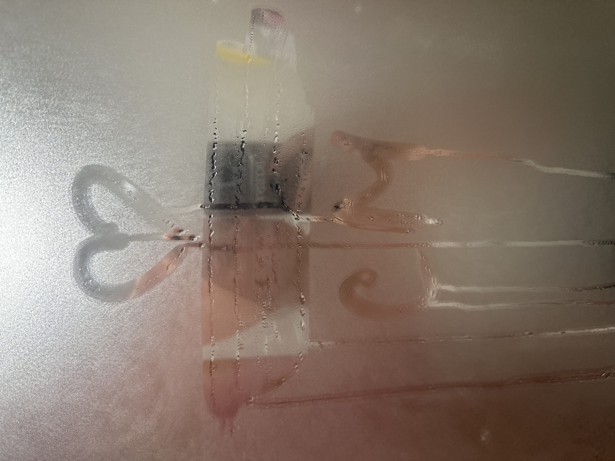 A close-up, slightly overexposed image shows a foggy shower door with the letters "ily" drawn on it. A shampoo or body wash bottle is visible behind the fog. The bottle is clear with black accents and a yellow spout, and it appears to be affixed to the shower wall via suction cups. There are water droplets on the glass. The fog obscures the background, but the shower stall appears to have light-colored tiles. There is no evidence to suggest that this image was taken in Votkinsk, Russia.