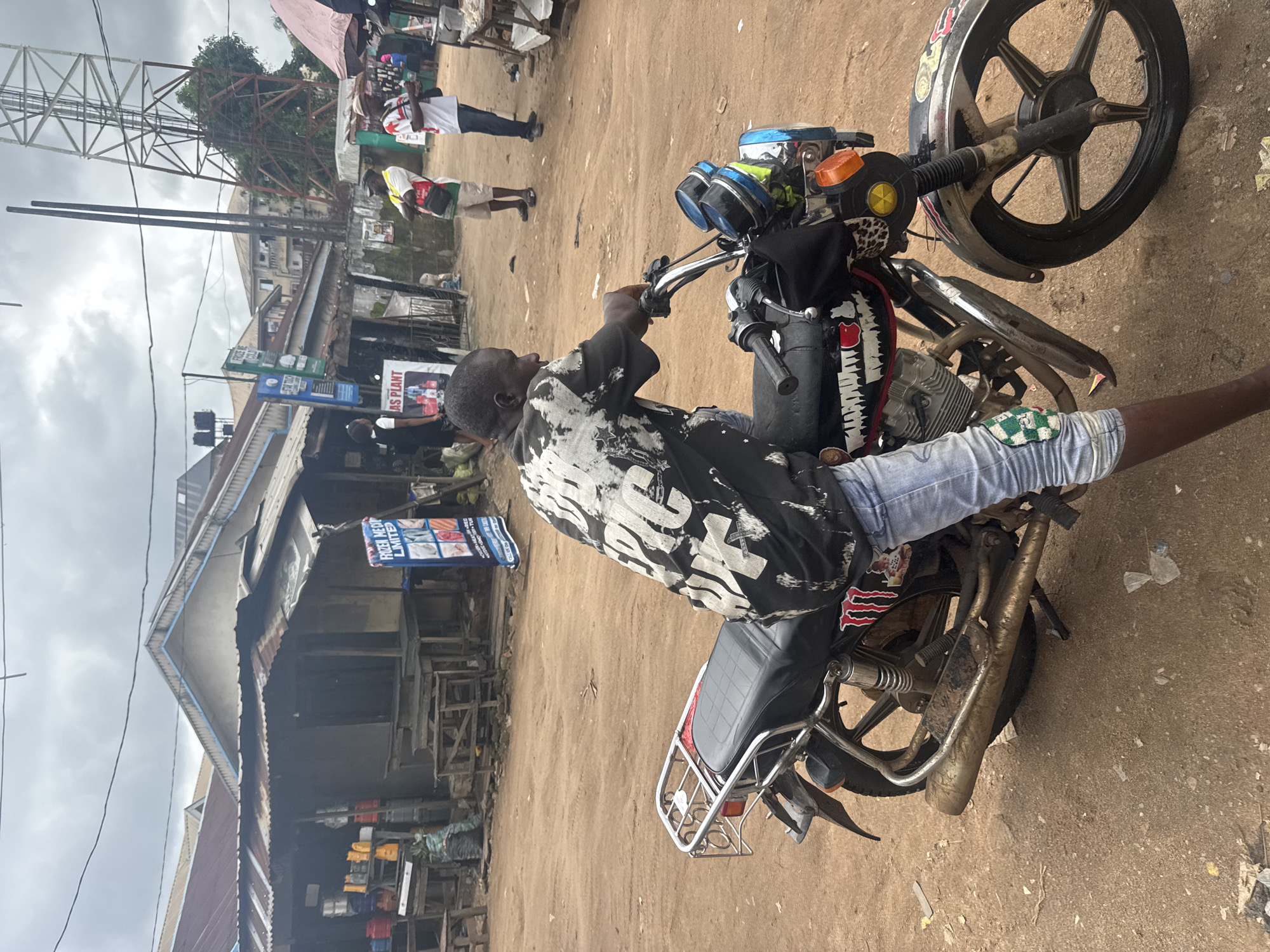 An outdoor, medium shot shows a man sitting on a motorcycle in what appears to be a marketplace in Ihiagwa, Nigeria. The man, wearing a black and white graphic t-shirt and light blue jeans with a green and white patched knee, has his hands on the motorcycle's handlebars. The motorcycle is black with red and white graphics on its fuel tank. The scene is dusty and unpaved, with several makeshift stalls and buildings lining the background. A tall telecommunications tower is visible in the upper right corner of the image. Several other individuals are present in the background, some standing and some walking, suggesting a busy commercial area. The sky is overcast with clouds.