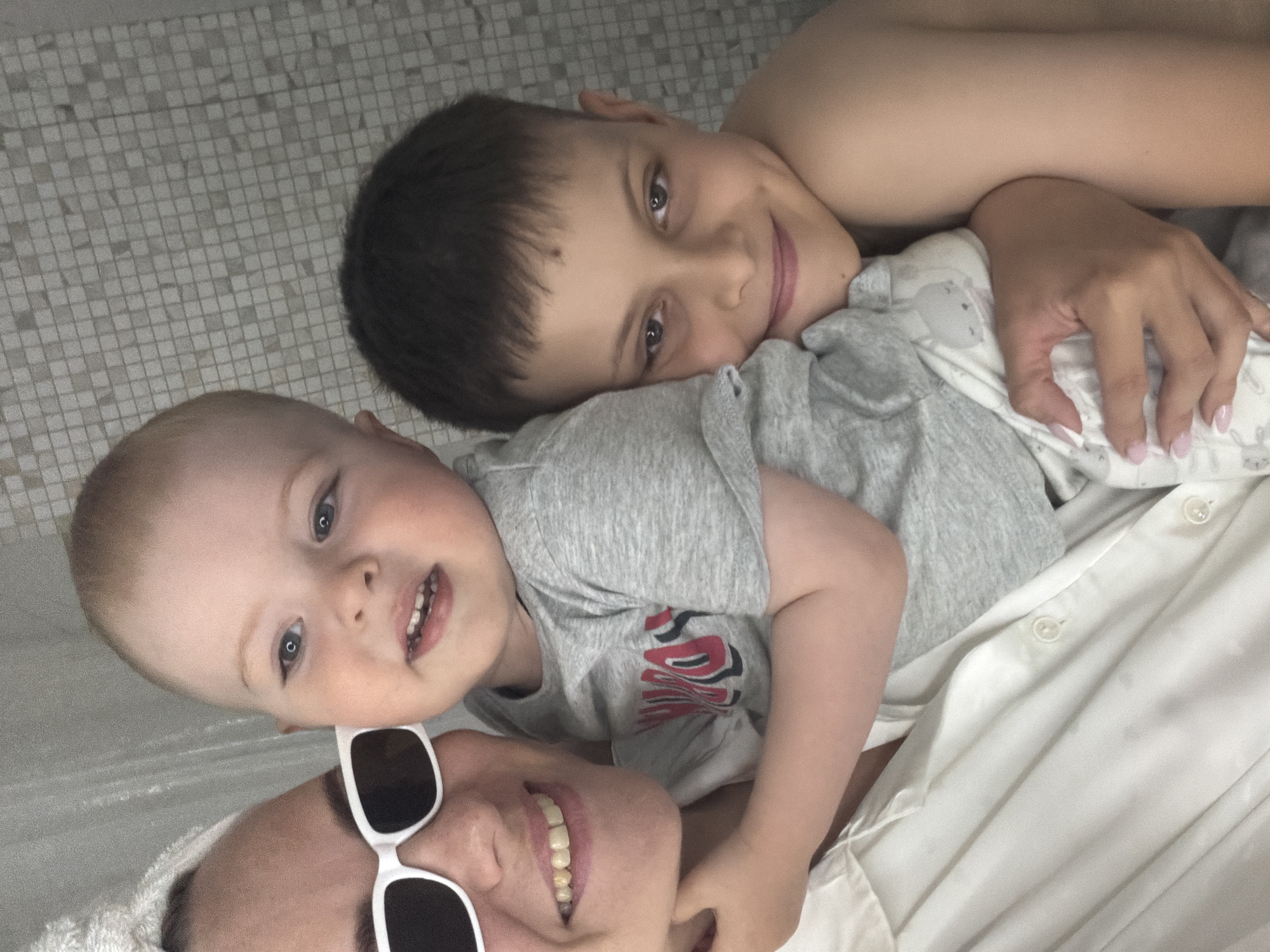 A family photo, consisting of a woman and two children, presumably her sons, lying down on a light-colored surface, possibly a bed or floor. The woman is wearing white sunglasses and a towel on her head. The younger child, wearing a gray t-shirt, is positioned between the woman and the older child, with the latter resting on the other side of the younger one. The older child is shirtless. The family members are interacting closely and appear to be happy and relaxed. The background shows a light-colored, patterned surface, possibly tiled, suggesting they are indoors. There is no evidence to indicate the city of Votkinsk, Russia.