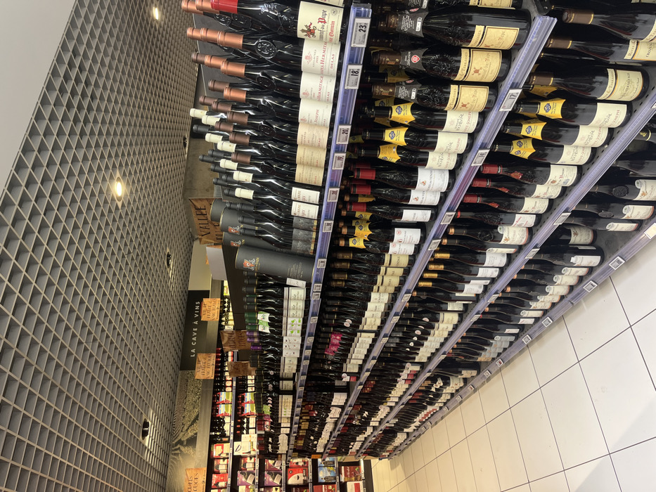 The image shows a wine aisle in a grocery store located at latitude 49.283 and longitude 3.999 in Saint-Brice-Courcelles, France. There are rows of wine bottles on metal shelves, organized by brand and type. The shelves are labeled with prices and information about the wines. The store has a white tiled floor and a white ceiling with black grid-style panels. The aisle is well lit. The sign above the shelves reads “La Cave à Vins” which translates to "The Wine Cellar". There are no visible people in the image. The wine bottles are all full and appear to be in good condition. The bottles are arranged in a neat and orderly fashion. There is no visible damage to the shelves or the bottles.