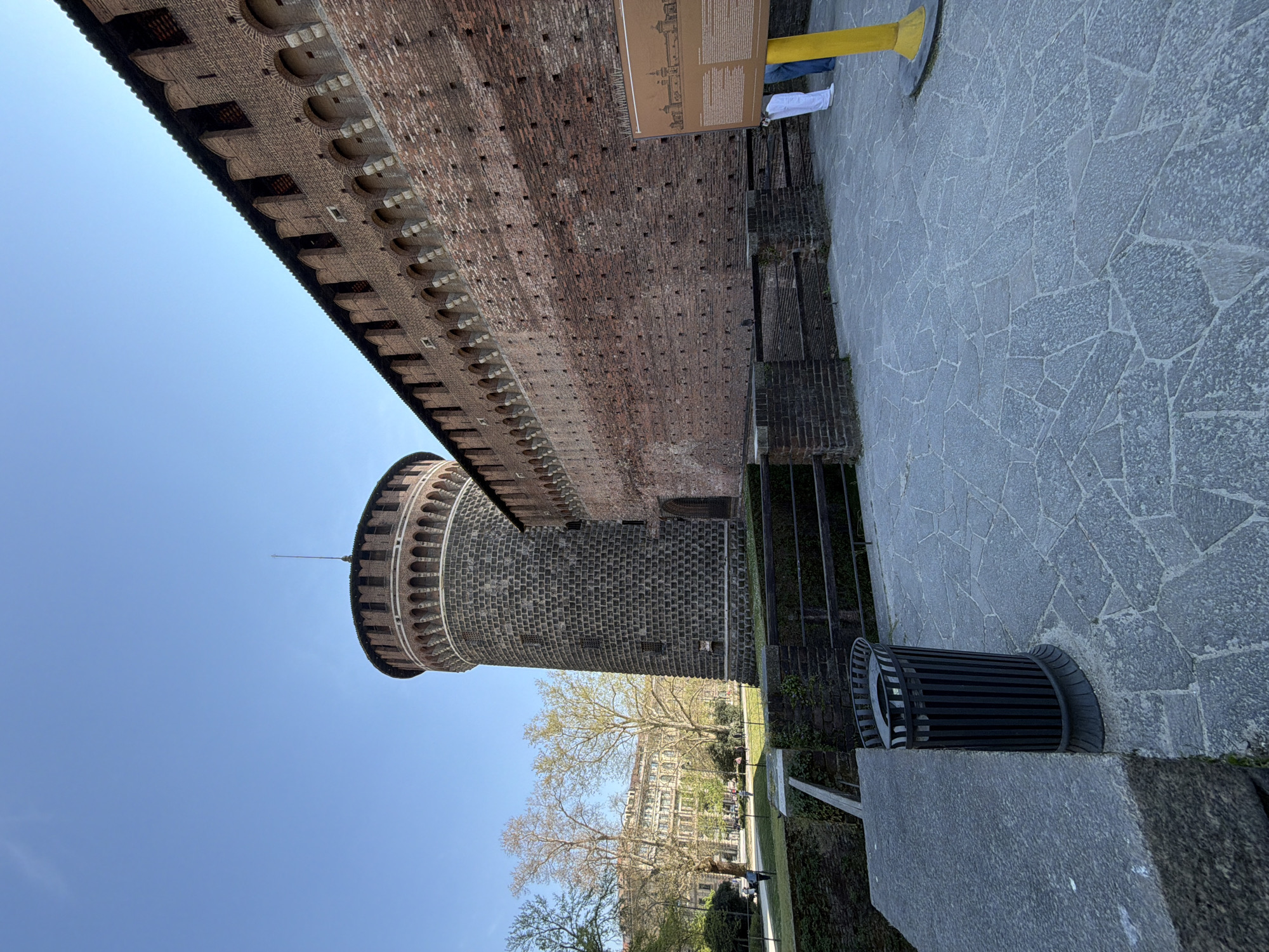 The image shows a low-angle view of Sforza Castle in Milan, Italy. A large brick wall with crenellations runs along the top. Attached to the wall is a cylindrical tower, also made of brick. The sky is clear and blue. A stone-paved area is visible on the right side of the image, along with a metal trash can. In the distance, trees and buildings can be seen beyond the castle walls. A sign with information about the castle is partially visible, with a person standing near it.