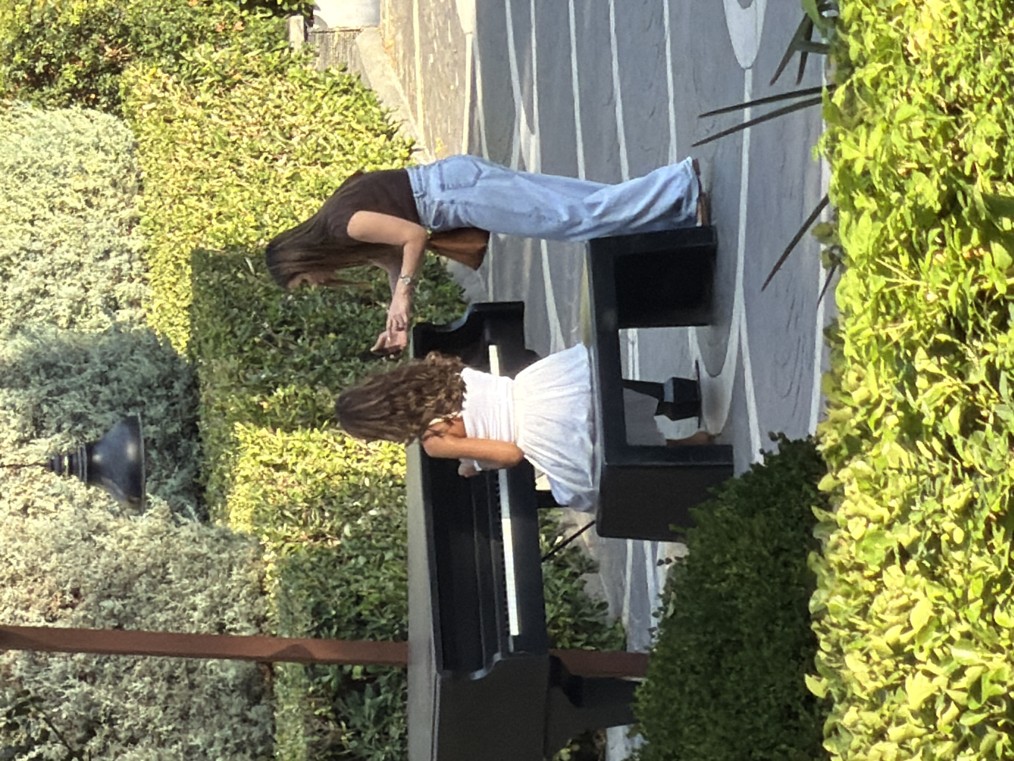 In Baku, Azerbaijan, a young girl with curly brown hair is seated at a black grand piano, wearing a white dress. She appears to be playing the piano. A woman with long brown hair, wearing a dark brown t-shirt and light blue wide-leg jeans, stands to the right of the piano, holding a smartphone in her hands and looking towards the girl. The scene takes place outdoors, surrounded by lush green bushes and trees. A lamppost with a dark shade is visible in the background. The ground is paved with a patterned surface. The overall atmosphere suggests a public space designed with natural elements.