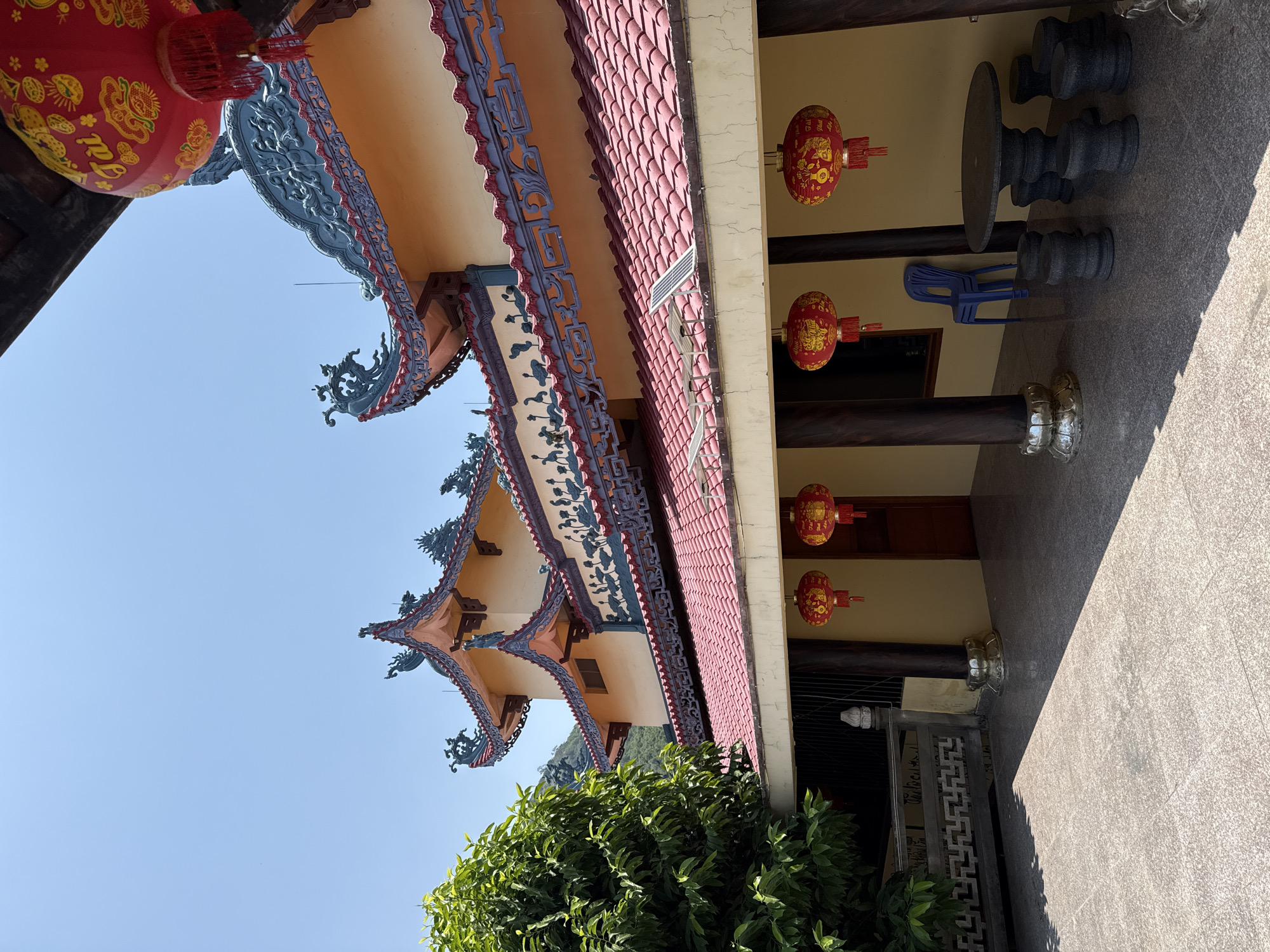 The image shows a portion of a building in Bắc Nha Trang, Vietnam, characterized by traditional East Asian architectural elements. Predominant features include layered, ornate roofs with red tiles and decorative blue and red trim. Red lanterns hang along a covered walkway supported by dark-colored columns. A stone table with stools is visible in the walkway, alongside a blue plastic chair. A small solar panel is mounted on the roof. The sky is clear and blue, and green foliage is visible near the base of the building. A decorative gate is also visible.