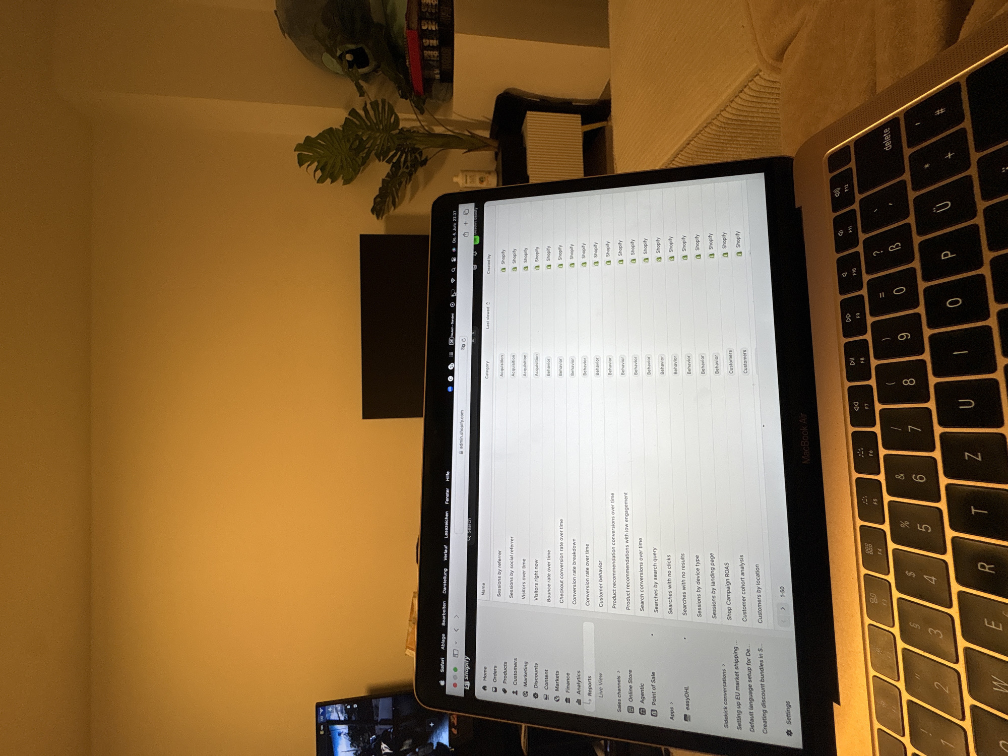 The image displays an indoor scene, likely a home office, captured in the late evening, as indicated by the time "Do. 4. Juni 22:37" (Thursday, June 4, 10:37 PM) visible on the laptop screen. No people are present.
The primary subject is an open MacBook Air laptop, positioned on a surface, with its screen displaying the Shopify administration interface at "admin.shopify.com" in a Safari browser. The screen shows a list of reports categorized by "Acquisition," "Behavior," and "Customers." Notable report titles include "Sessions by referrer," "Visitors over time," "Bounce rate over time," "Checkout conversion rate over time," "Customer behavior," "Product recommendation conversions over time," and various "Searches" reports (by search query, with no clicks, with no results). The left sidebar of the Shopify interface shows navigation options such as "Home," "Orders," "Products," "Customers," "Marketing," "Analytics," and "Reports."
Part of the MacBook Air's keyboard is visible on the right, featuring a German layout with keys like 'Ü', 'Ö', 'Ä', and the swapped 'Z' and 'Y' keys, which aligns with the location context of Mönchengladbach, Germany.
In the background, a green Monstera plant adds a touch of greenery. A dark monitor or television screen, a white ventilation grille or speaker, and a light-colored sofa or armchair are also partially visible. The walls are a plain, warm yellow hue, suggesting artificial indoor lighting. The scene conveys a sense of someone working on e-commerce management late at night.