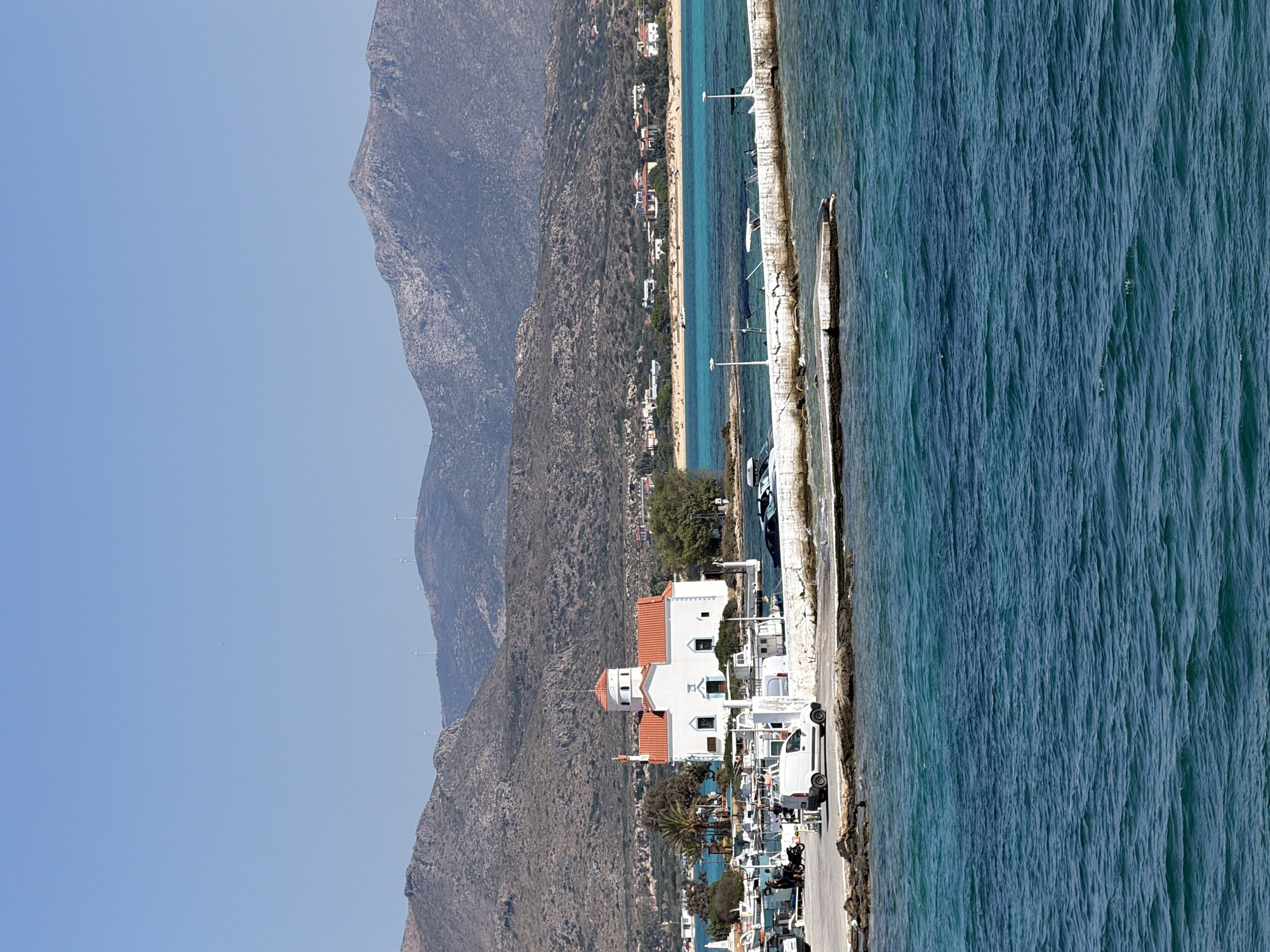 In Elafonisos, Greece, a white church with a red tiled roof is situated on a small island or peninsula connected to the mainland by a concrete structure. Several boats, including fishing boats and smaller vessels, are docked along the shore. A white van is parked on the concrete area. In the background, a coastal town with white buildings and red roofs lines a beach, with a backdrop of dry, mountainous terrain under a clear blue sky. Wind turbines are visible on the distant hills. The foreground features the turquoise waters of the sea with visible waves.