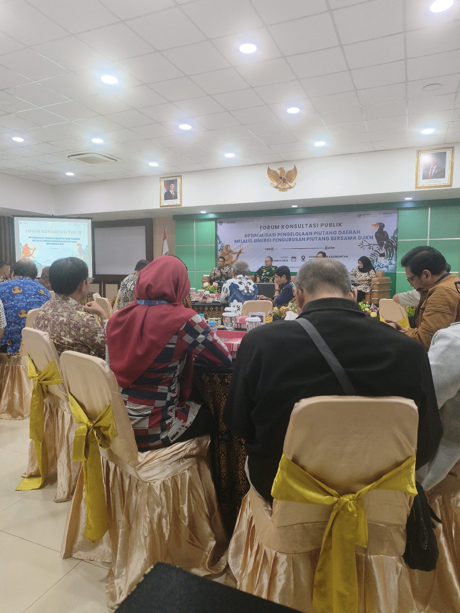 The image depicts an indoor conference or seminar setting, possibly at the Kanwil DJKN Kalimantan Timur in Samarinda, Indonesia. The room features a white tiled floor and a drop ceiling with recessed lighting. A projector screen displaying "FORUM KONSULTASI PUBLIK OPTIMALISASI PENGELOLAAN PIUTANG DAERAH MELALUI SINERGI PENGURUSAN PIUTANG BERSAMA DJKN" is visible on the left. The room is decorated with banners and portraits on the walls, including a prominent Indonesian coat of arms.
In the foreground and midground, several people are seated at tables, facing a stage area where a speaker is addressing the audience. The attendees are seated in chairs covered with gold fabric and adorned with yellow sashes tied in bows. The individuals are dressed in formal or semi-formal attire. Some are facing the stage, while others are engaged in conversation or looking at their phones. The overall atmosphere suggests a professional gathering.
