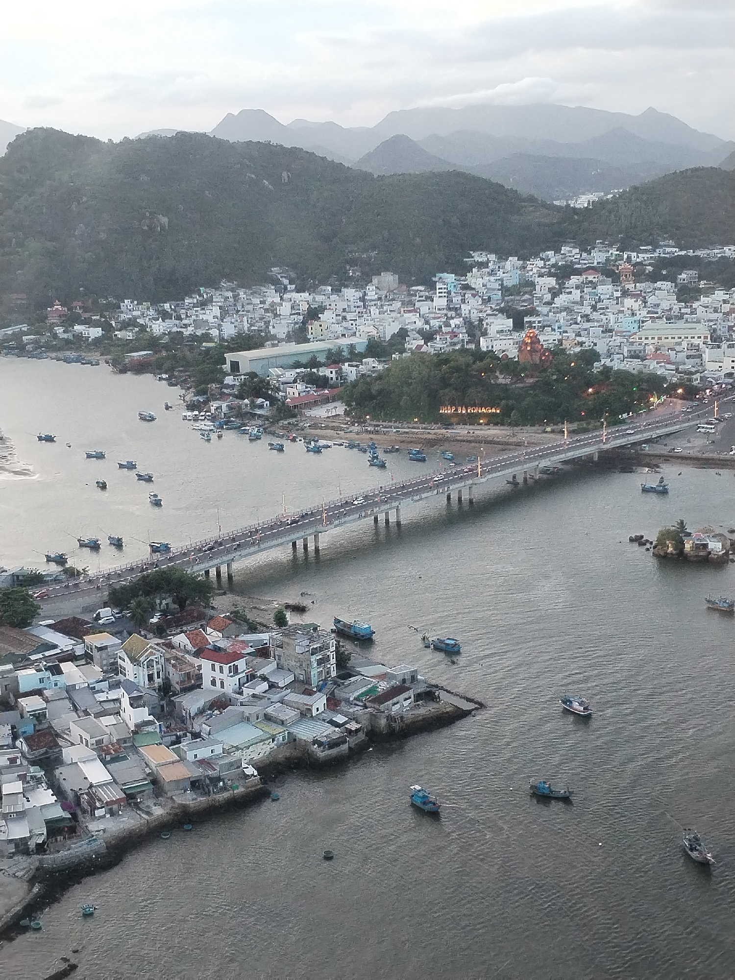 The image depicts a cityscape in Nha Trang Ward, Vietnam, with a focus on a bridge spanning a waterway. Mountains form a backdrop to a dense urban area characterized by closely packed buildings, primarily white with occasional colorful roofs. A bridge with multiple support pillars crosses the water, carrying vehicular traffic. Numerous boats are scattered across the water's surface, and a smaller coastal area is visible in the foreground with low-lying buildings. The sky is overcast, suggesting an evening or early morning time.