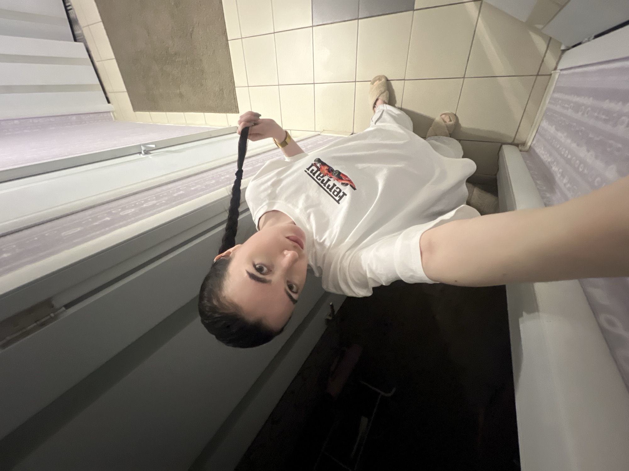 A young person is lying on a tanning bed, taking a selfie. The person is wearing a white t-shirt with a red and black Ferrari logo on it, and fuzzy slippers. Their hair is pulled back into a long braid. The setting appears to be a tanning salon, with the tiled walls and tanning bed clearly visible. The lighting suggests it is indoors, and the time of day is not discernible. The person's facial expression is neutral, and they are holding their braid with one hand. The image is taken from a high angle, looking down at the person.
