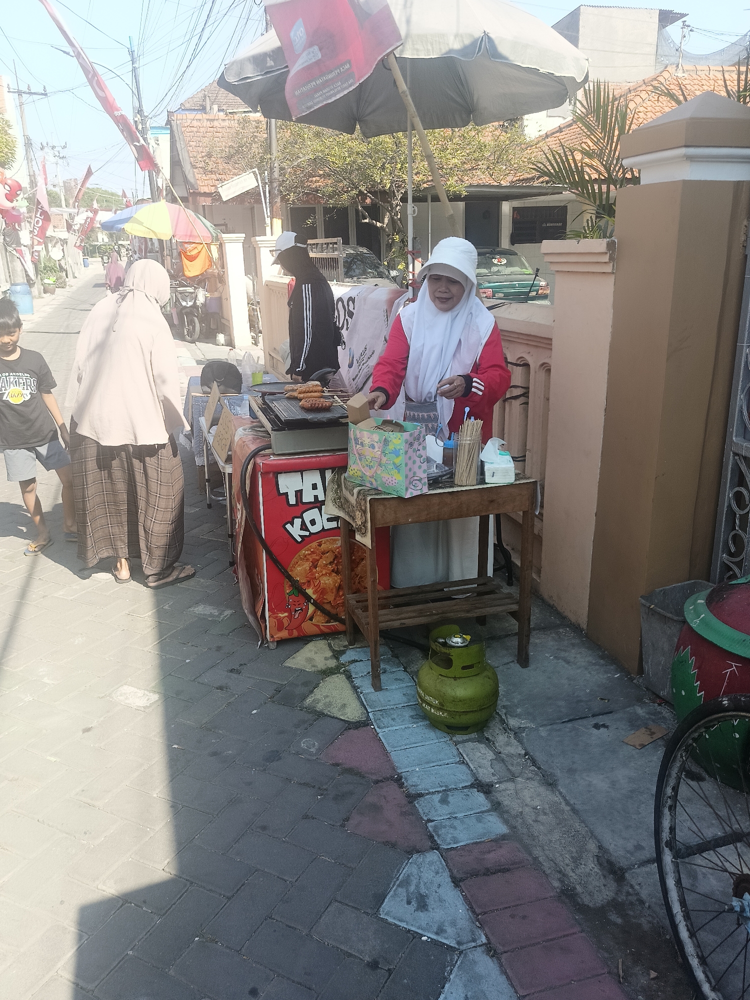 In the city of Gedangan, Indonesia, on a sunny day, a woman wearing a white hijab and a red jacket is tending to a street food stall. The stall features a grill with several sausages cooking and a red cooler adorned with text and an image of chilies. Beside the stall, a green gas cylinder is visible on the ground. Other people, including a child in a black Lakers t-shirt and a person in a beige hijab and checkered pants, are present near the stall. In the background, a street lined with buildings and trees can be seen, with flags and banners adding to the vibrant atmosphere. The ground is paved with interlocking bricks, and a bicycle wheel is partially visible on the right side of the image.