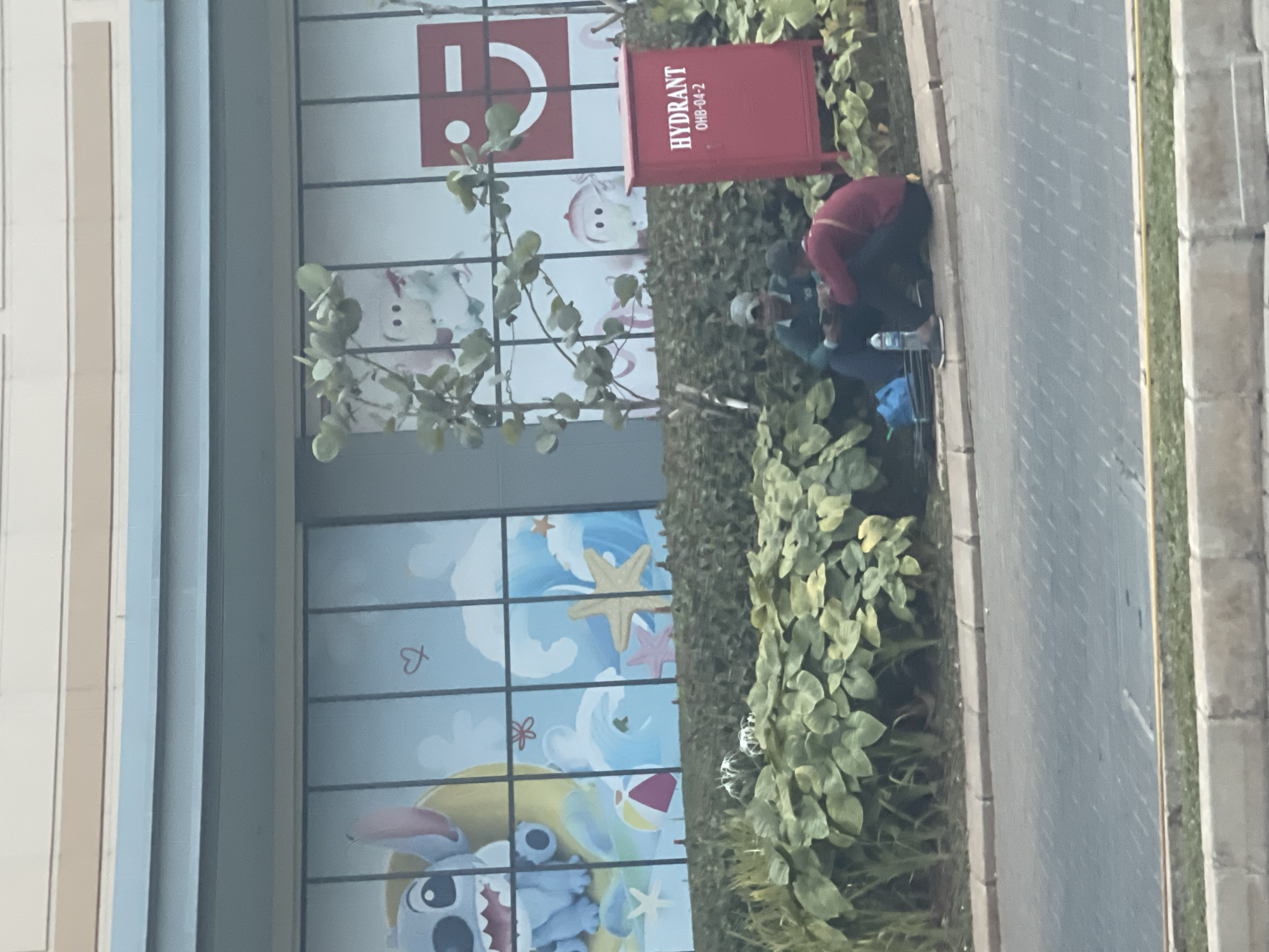 Two individuals are seated on the ground near a red hydrant box, with a large, leafy green bush behind them. The individual on the left, wearing a dark green shirt and a grey cap, is looking down at a mobile phone. The individual on the right, wearing a dark red shirt, is facing the other person, appearing to be engaged in conversation or interaction. A blue bag and a water bottle are on the ground near them. In the background, large glass panels display colorful murals, one featuring cartoon characters like Stitch and another with abstract designs and what appears to be a smiley face logo. The scene takes place outdoors on a paved area next to a street. The presence of a hydrant suggests an urban or commercial setting. This scene is likely located in the city of Bekasi, Indonesia, as indicated by the provided context.