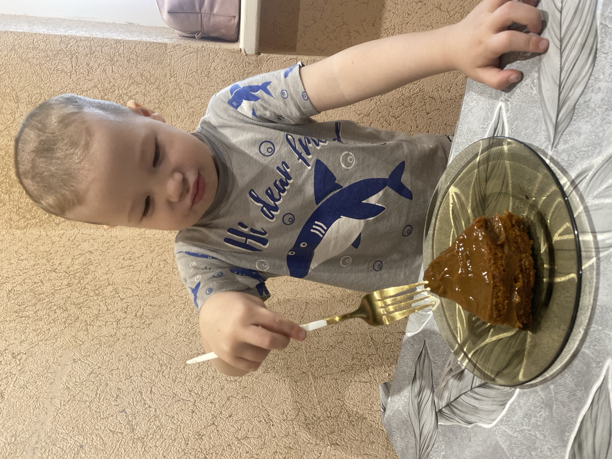 A child is about to eat a piece of cake with a fork. The child is wearing a gray t-shirt with a shark pattern. The cake is on a dark glass plate with a thin gold border. The plate sits on a table with a gray and white leaf pattern tablecloth. The wall in the background is textured and tan. The location is implied to be in Sochi, Russia, but no specific elements in the image corroborate the geographical location.