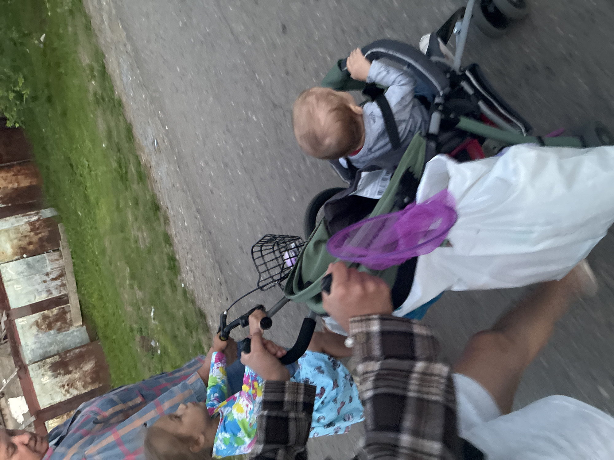 The image depicts a daytime outdoor scene on a dirt path, likely in Gornye Kljuchi, Russia. In the foreground, two individuals are partially visible, one pushing a stroller and another holding onto its handlebar. The stroller contains a baby wearing a gray shirt and appears to have a purple net bag and a white plastic bag attached. Behind the stroller, a young girl in a colorful, tie-dye shirt and a blue dress is walking, holding the handlebar of what seems to be a bicycle. The background shows a grassy area next to a row of dilapidated garages or sheds with rusted metal doors, some bearing numbers. The overall impression is a family outing or errand.